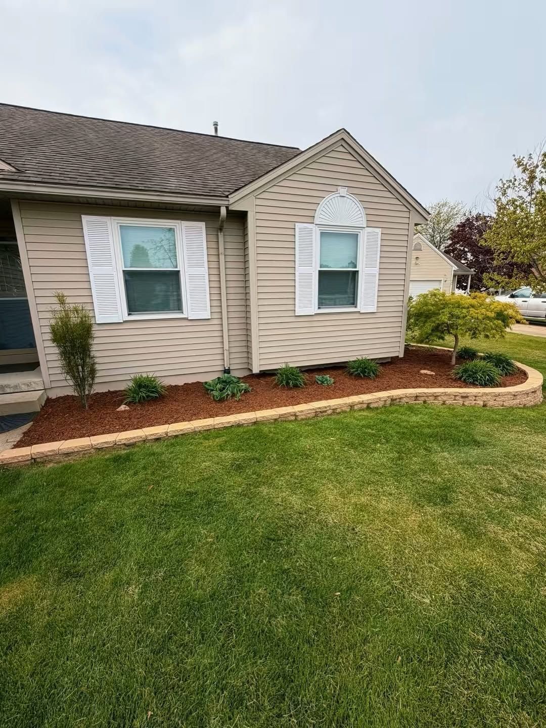Single-story beige house with white shutters, green lawn, and mulched flower bed along the front facade
