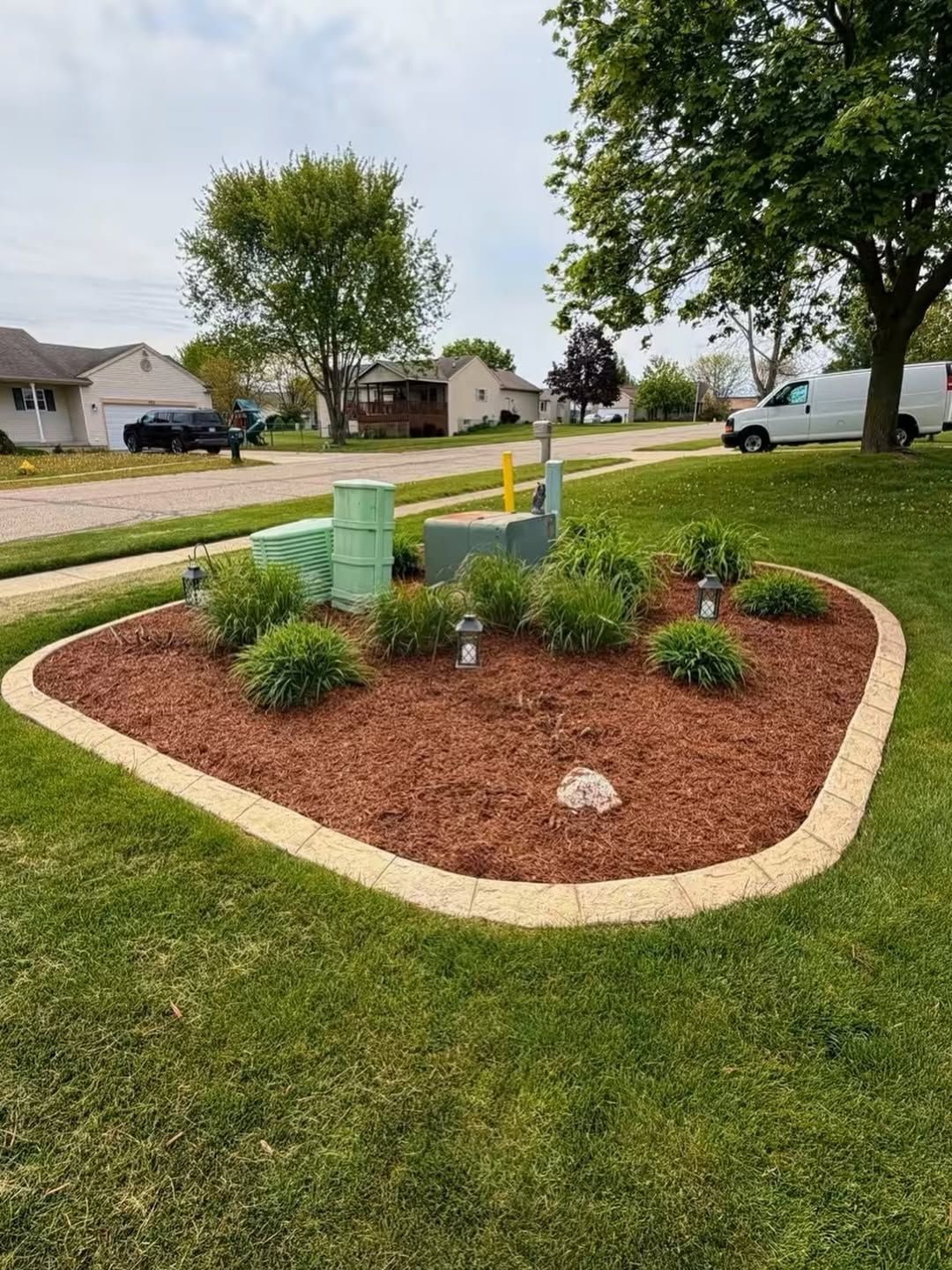 Small landscaped island with mulch, shrubs, utility boxes, and a yellow post in a suburban neighborhood.