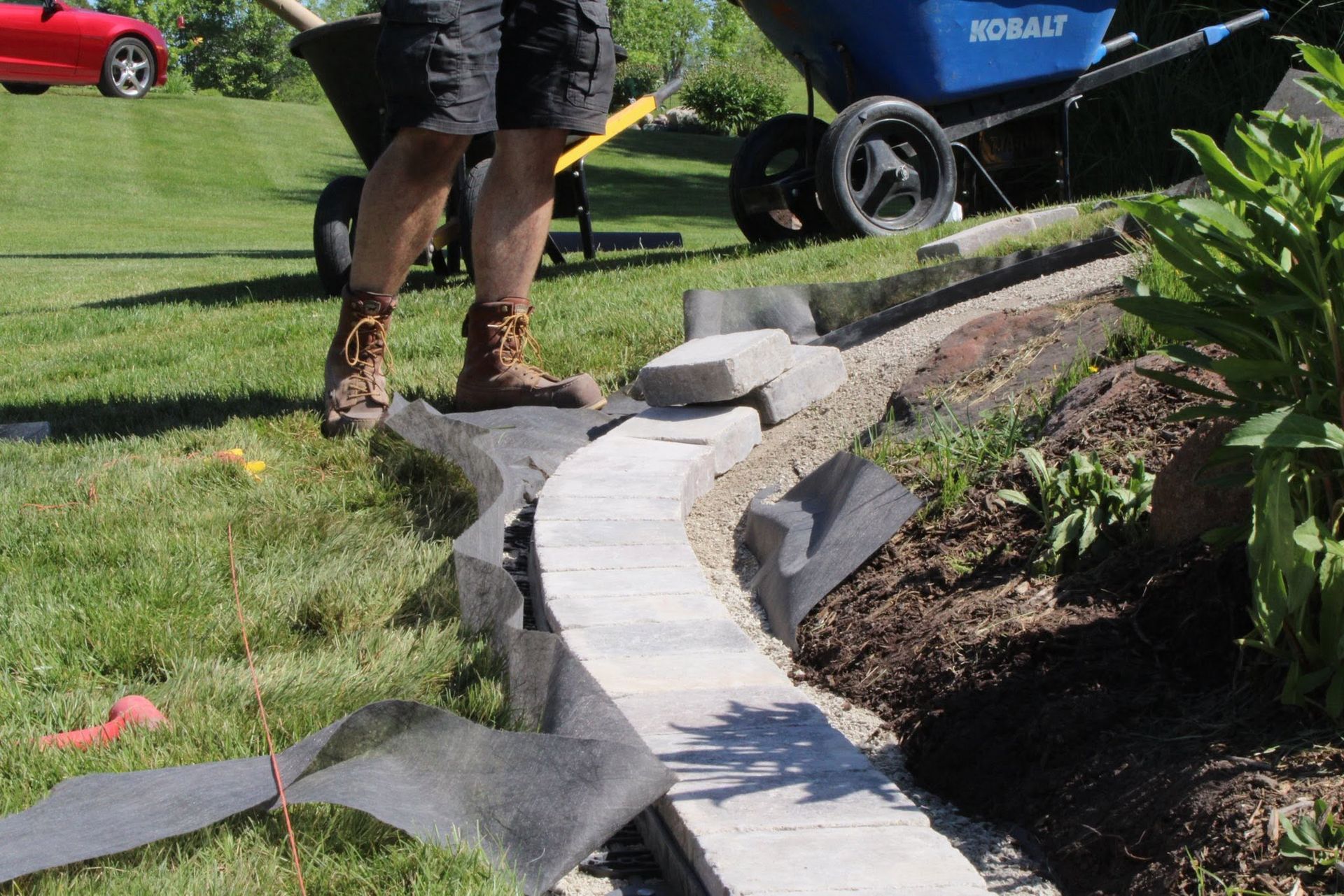 Worker installing curved stone edging along a garden path near a lawn and wheelbarrow.