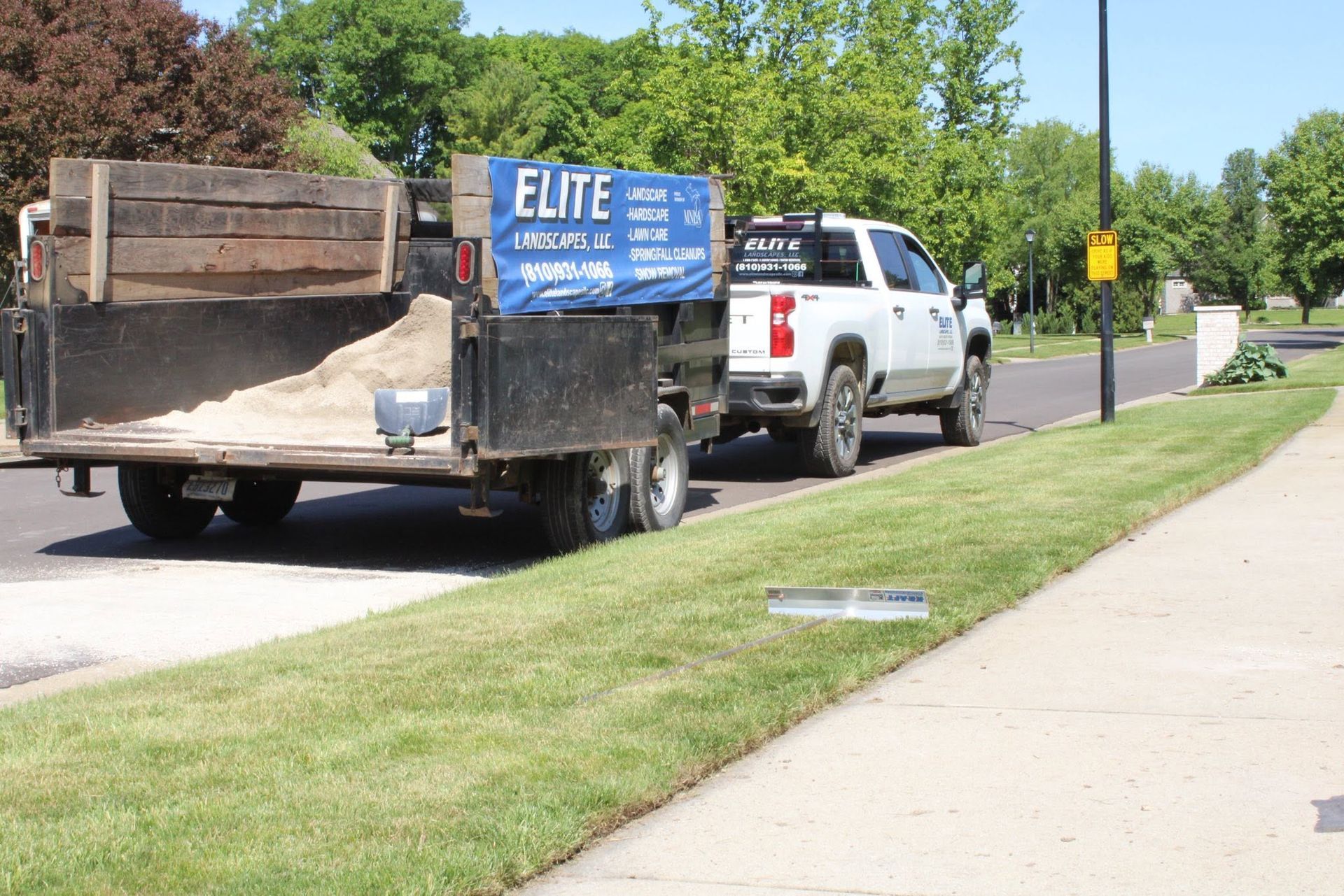 White pickup towing a blue utility trailer parked by a sidewalk on a sunny suburban street