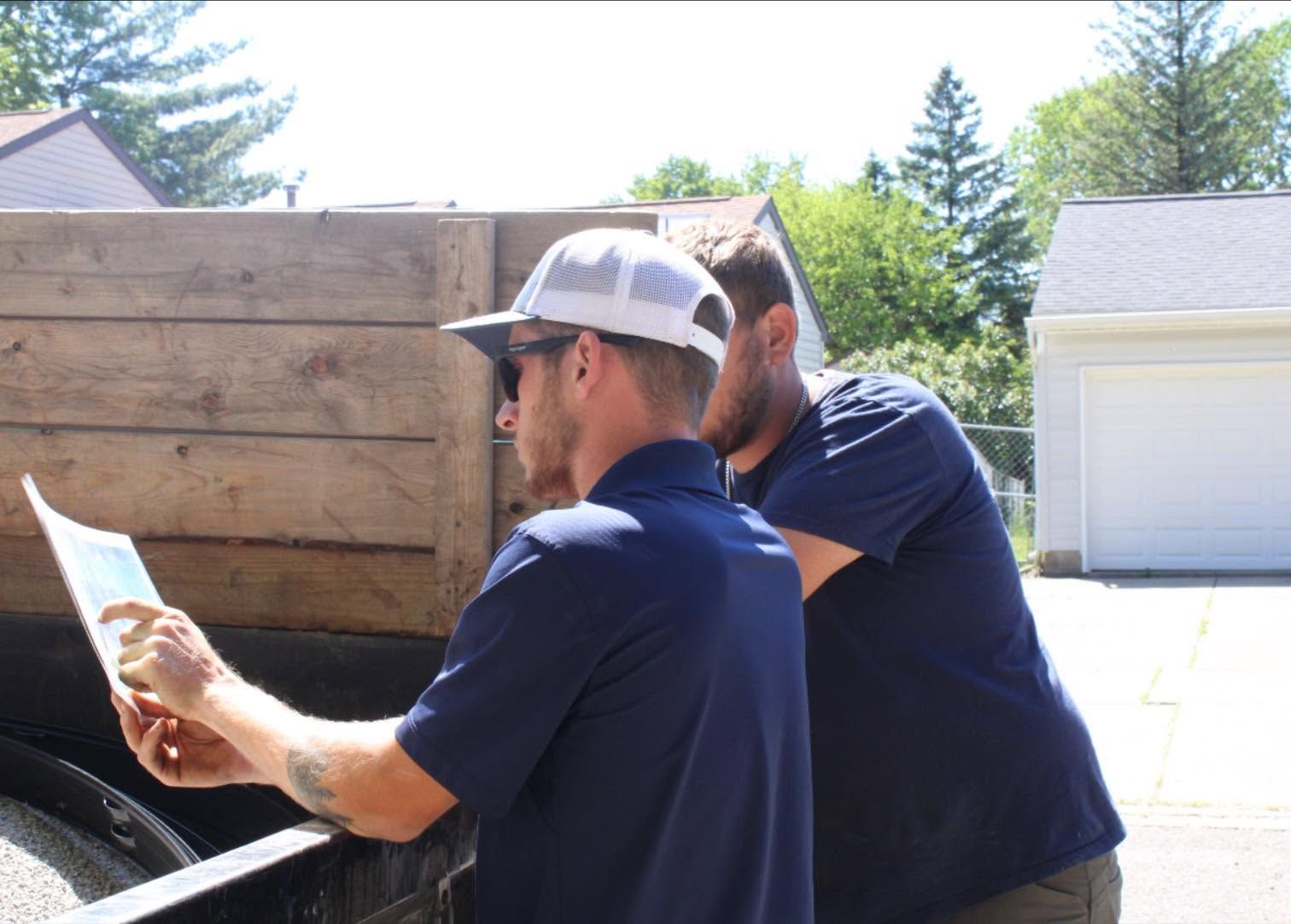 Two men in baseball caps sanding a wooden surface outdoors in bright sunlight