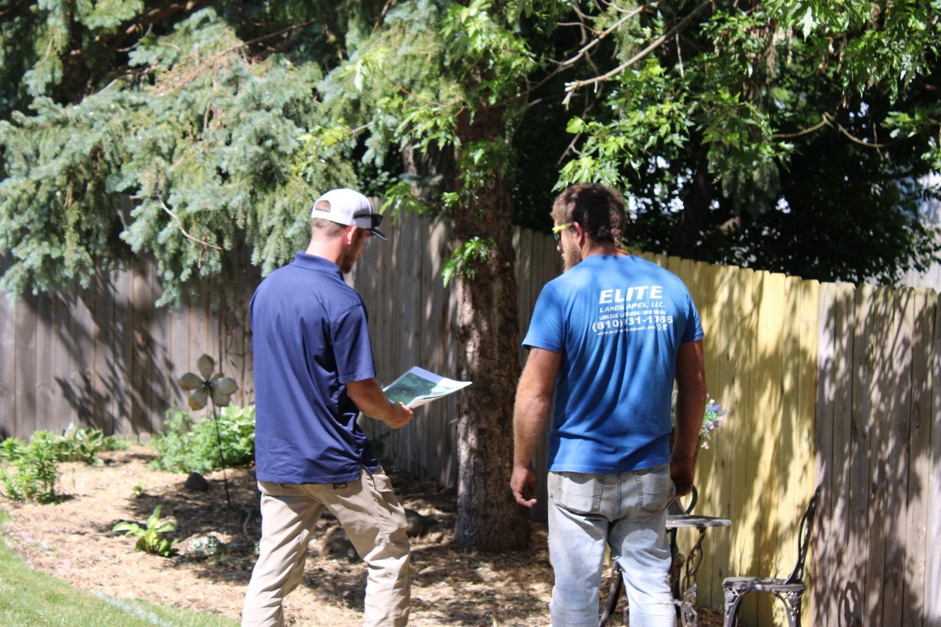 Two men inspect a fenced backyard, one holding a clipboard and both facing a tree.