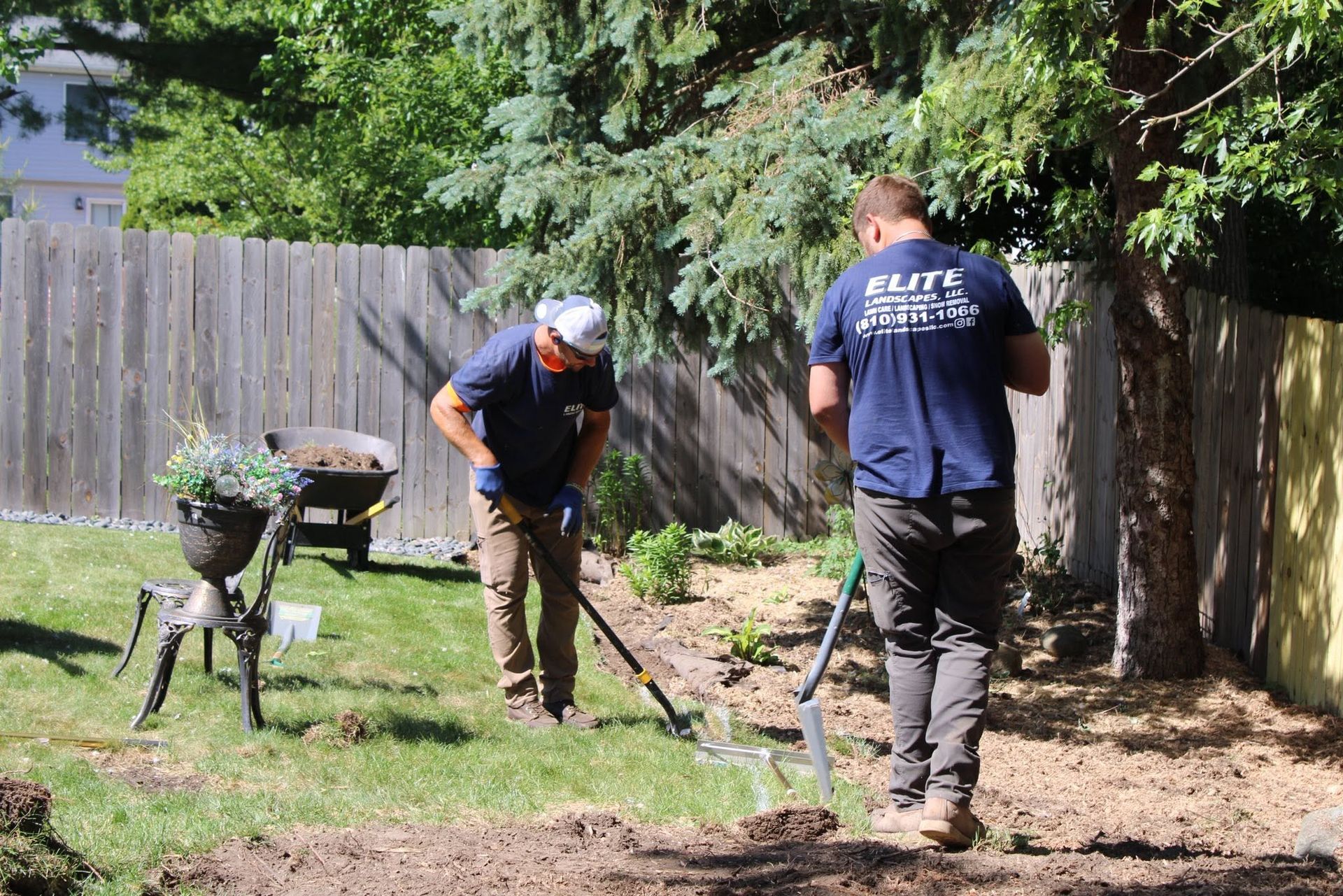 Two workers raking dirt in a backyard beside a wheelbarrow and wooden fence