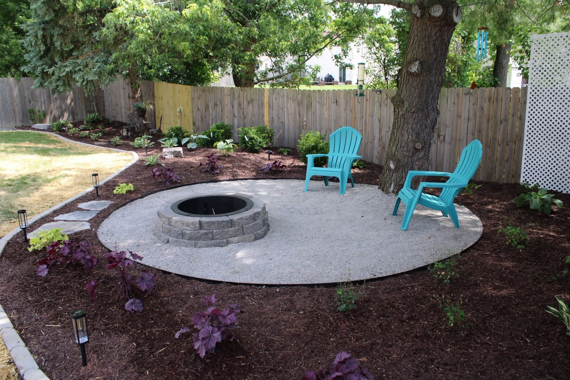 Backyard fire pit area with two turquoise chairs on a circular gravel patio, surrounded by mulch and a fence