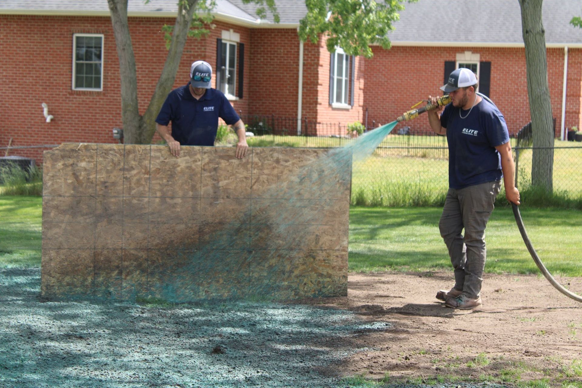 Two workers spraying water at a large outdoor surface in a yard beside a brick building