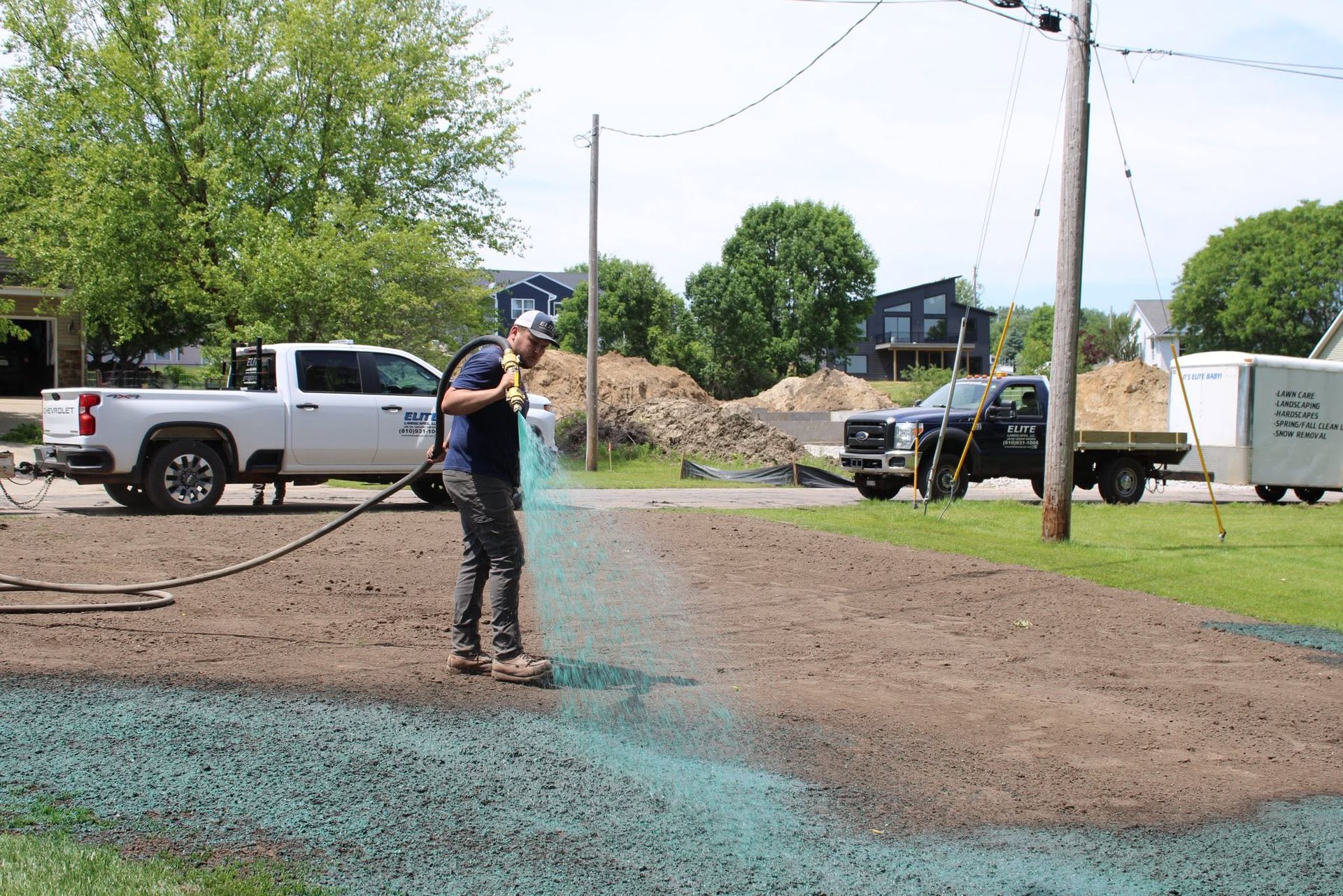 Worker pressure-washing a muddy driveway beside parked trucks and a utility pole in a rural yard