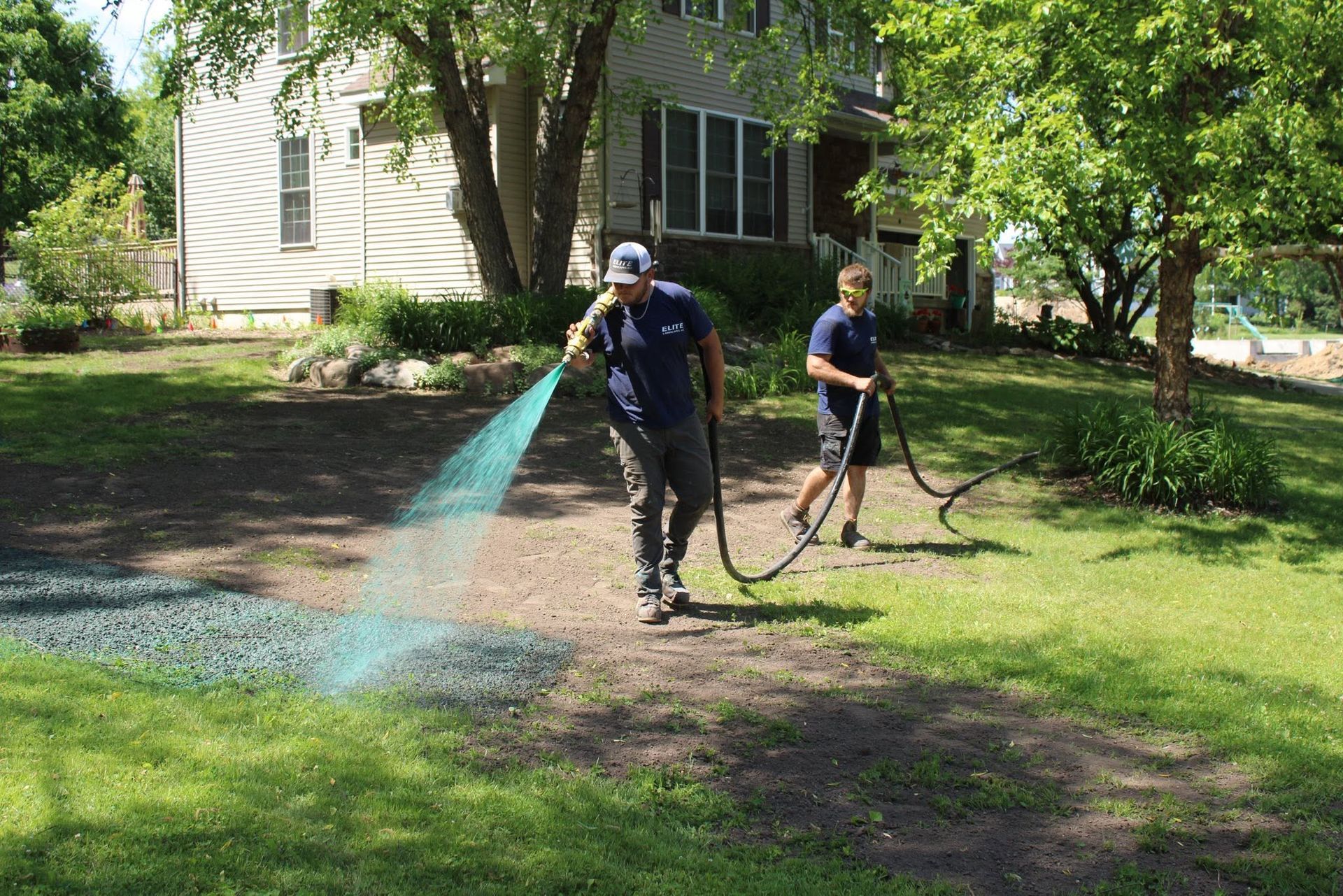 Two workers spraying water on a lawn beside a house in a sunny yard