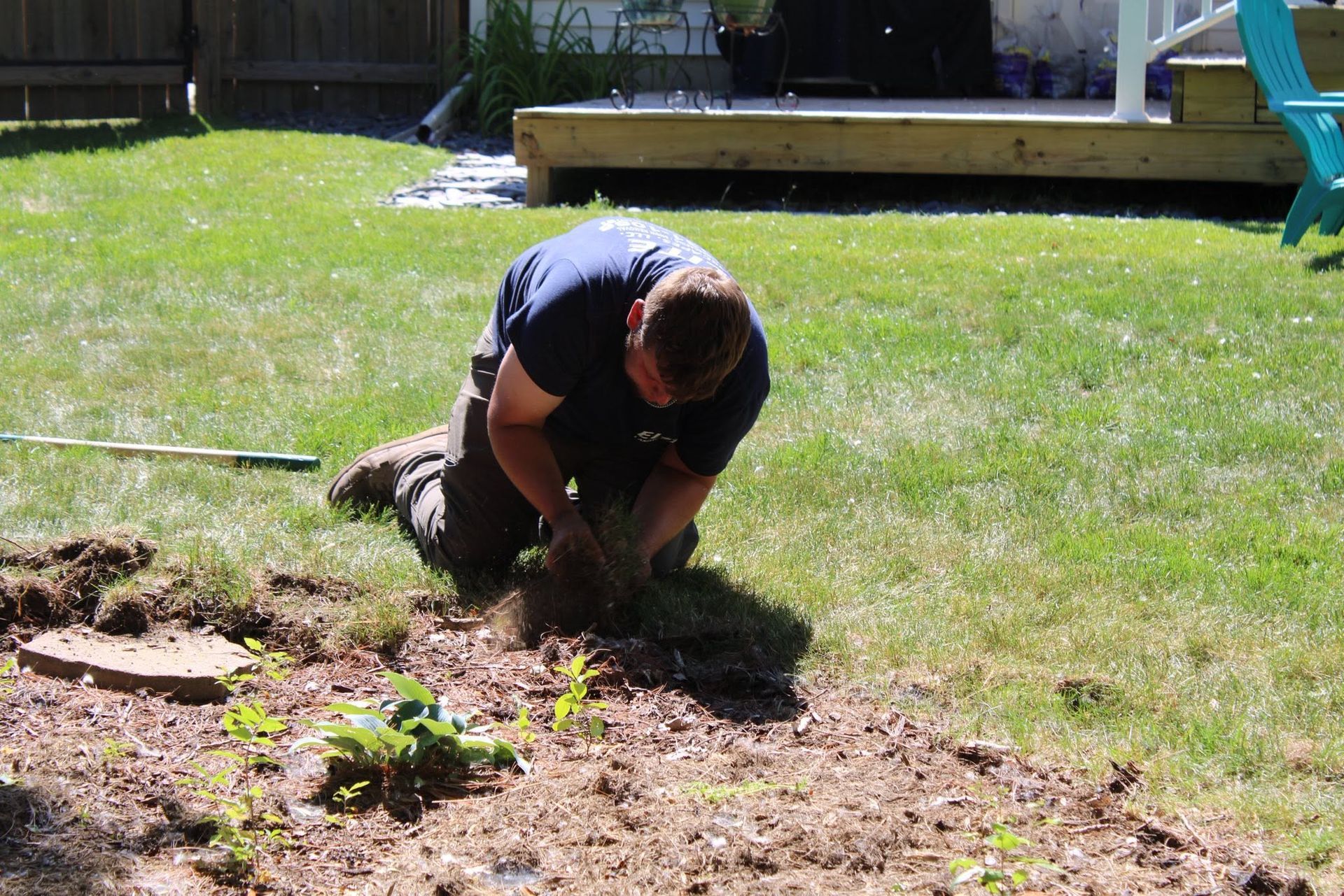 Person kneeling in a sunny backyard, digging in a patch of dirt beside the lawn.