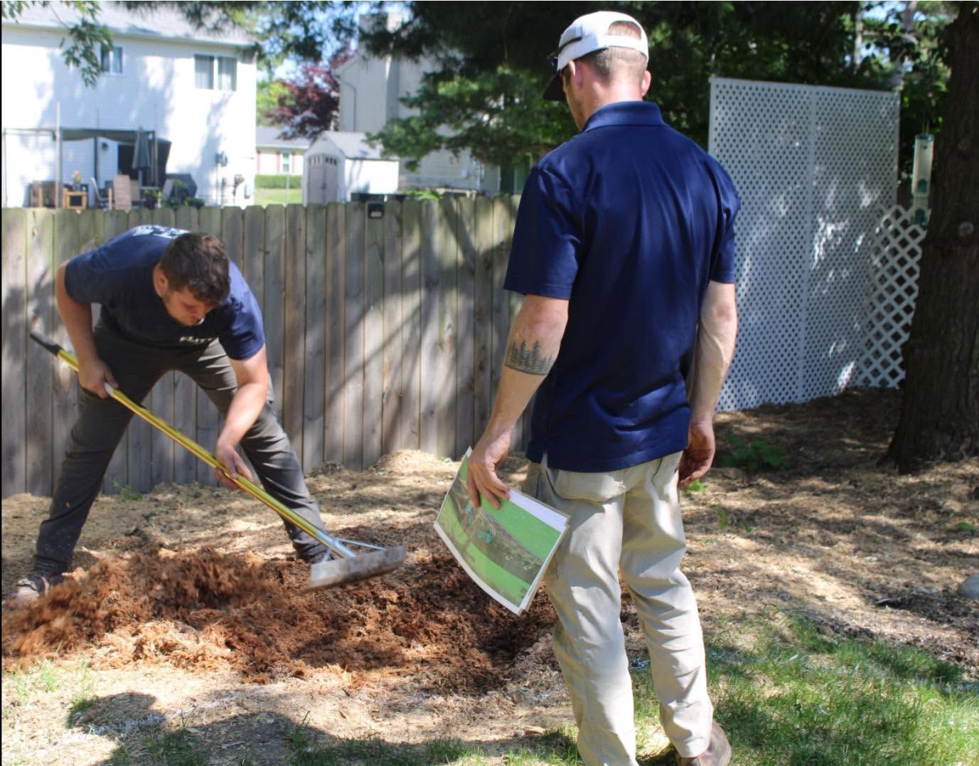 Two men work in a yard, raking mulch near a wooden fence and lattice gate.