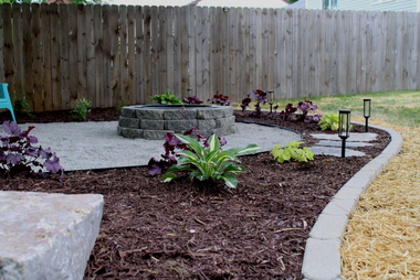 Curved backyard garden bed with mulch, hostas, stone edging, and a wooden fence.