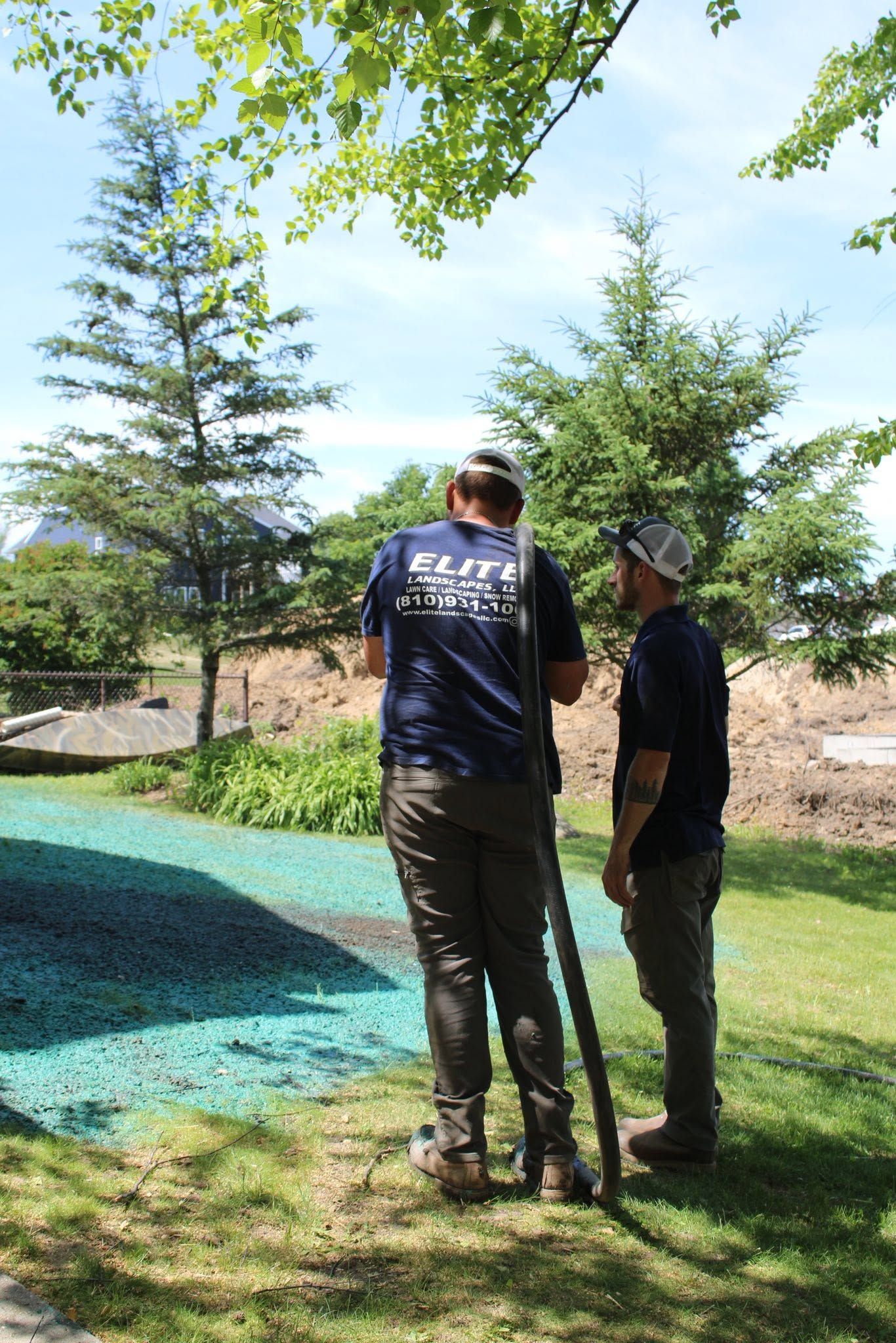 Two people stand on a grassy lawn beside a pond, under leafy trees on a sunny day.