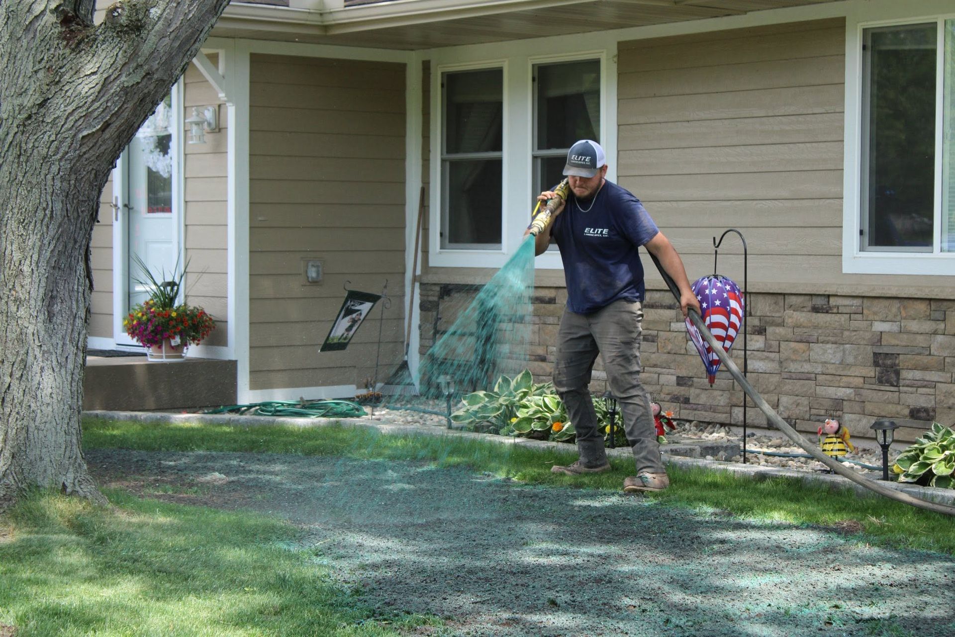 Person watering a lawn with a hose beside a house and sidewalk in a yard