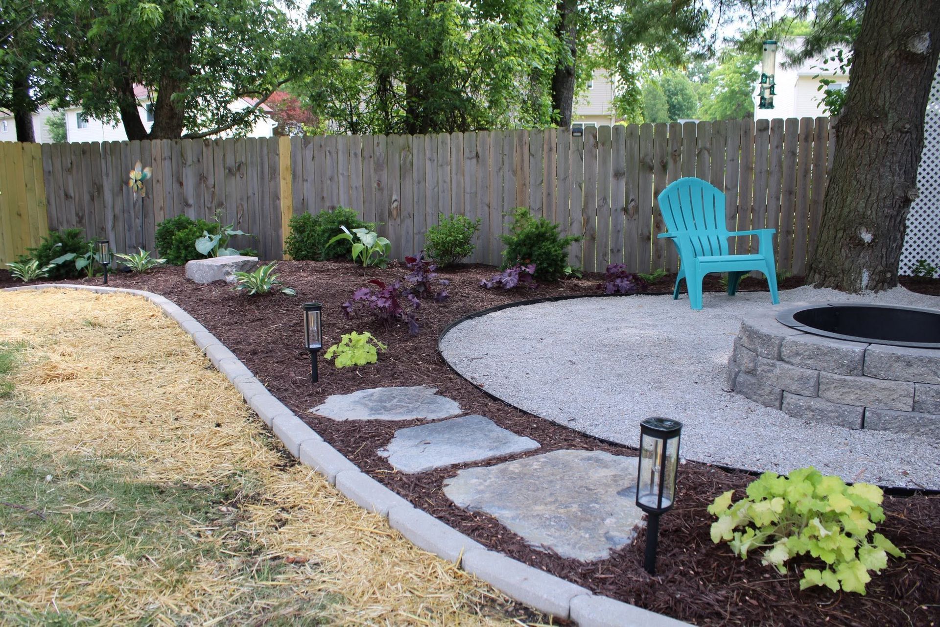 Backyard garden with curved stone path, mulch beds, tree, and turquoise chair beside a fire pit