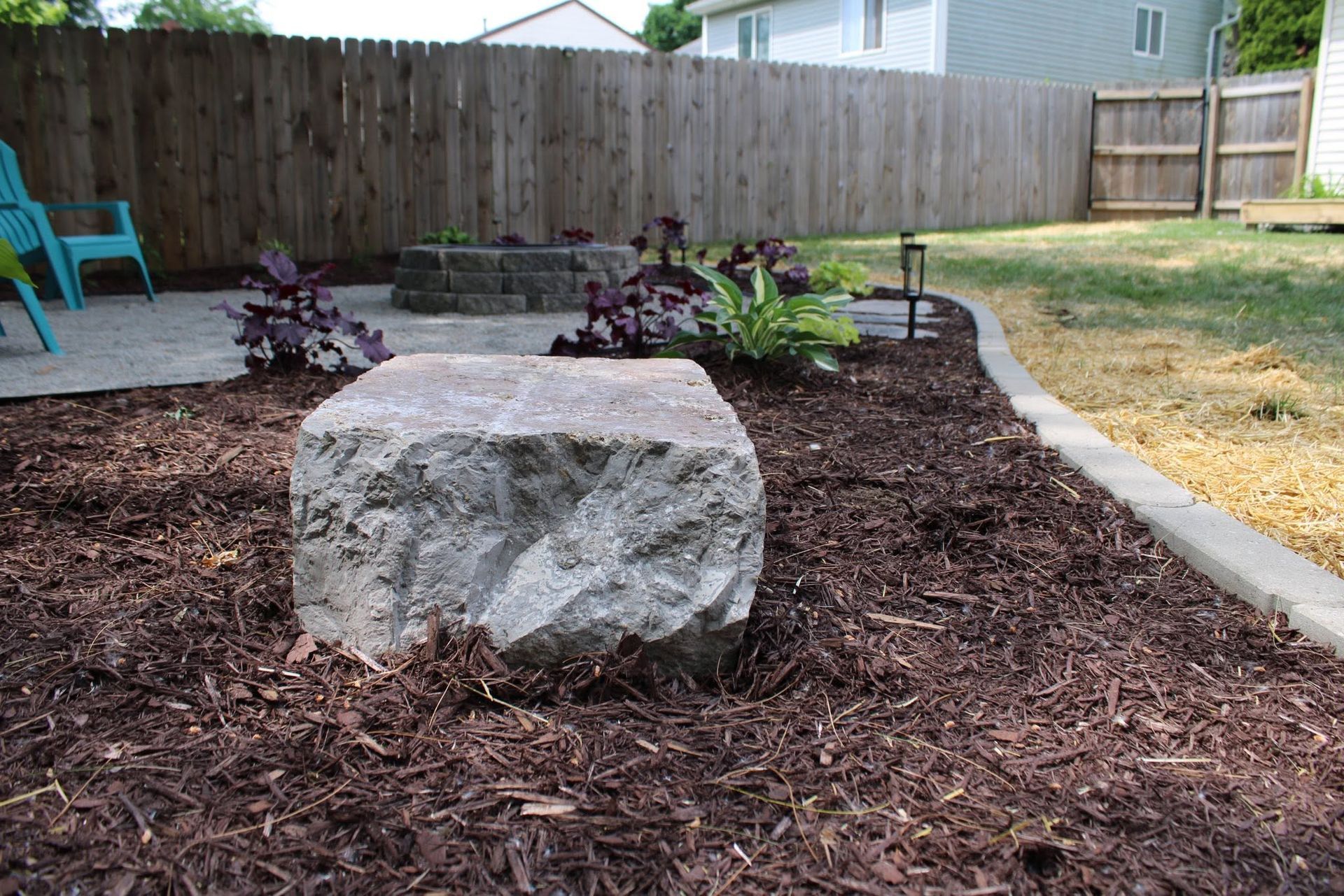 Curved mulch garden bed with potted plants, black chairs, and solar lights beside a fence and lawn.
