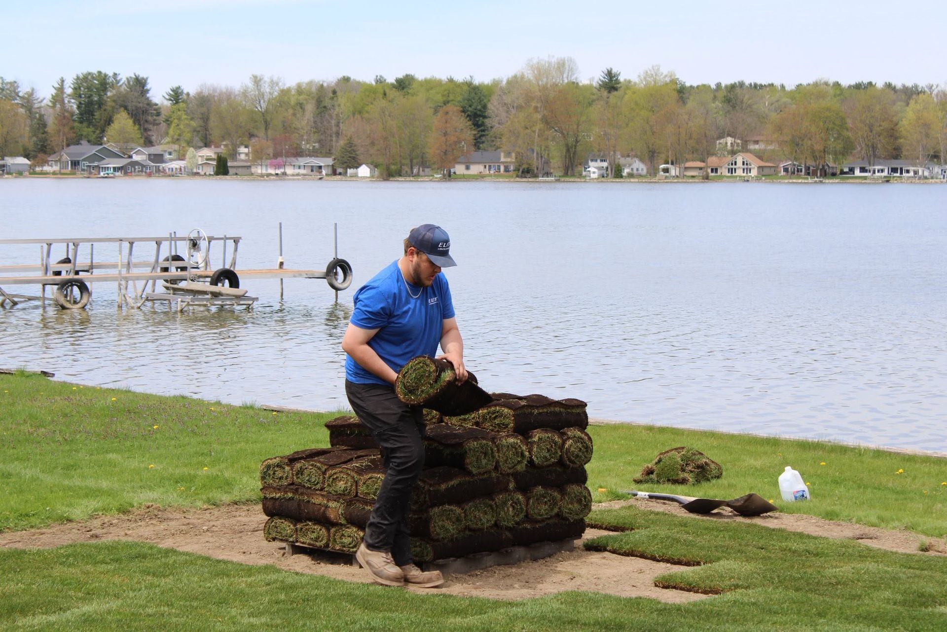 Person stacking moss-covered stones by a lakeshore with docks and houses in the background