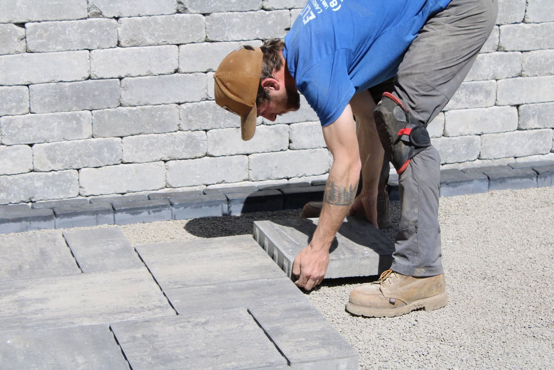 Worker in blue shirt and tan cap laying pavers on a paved surface beside a cinderblock wall.