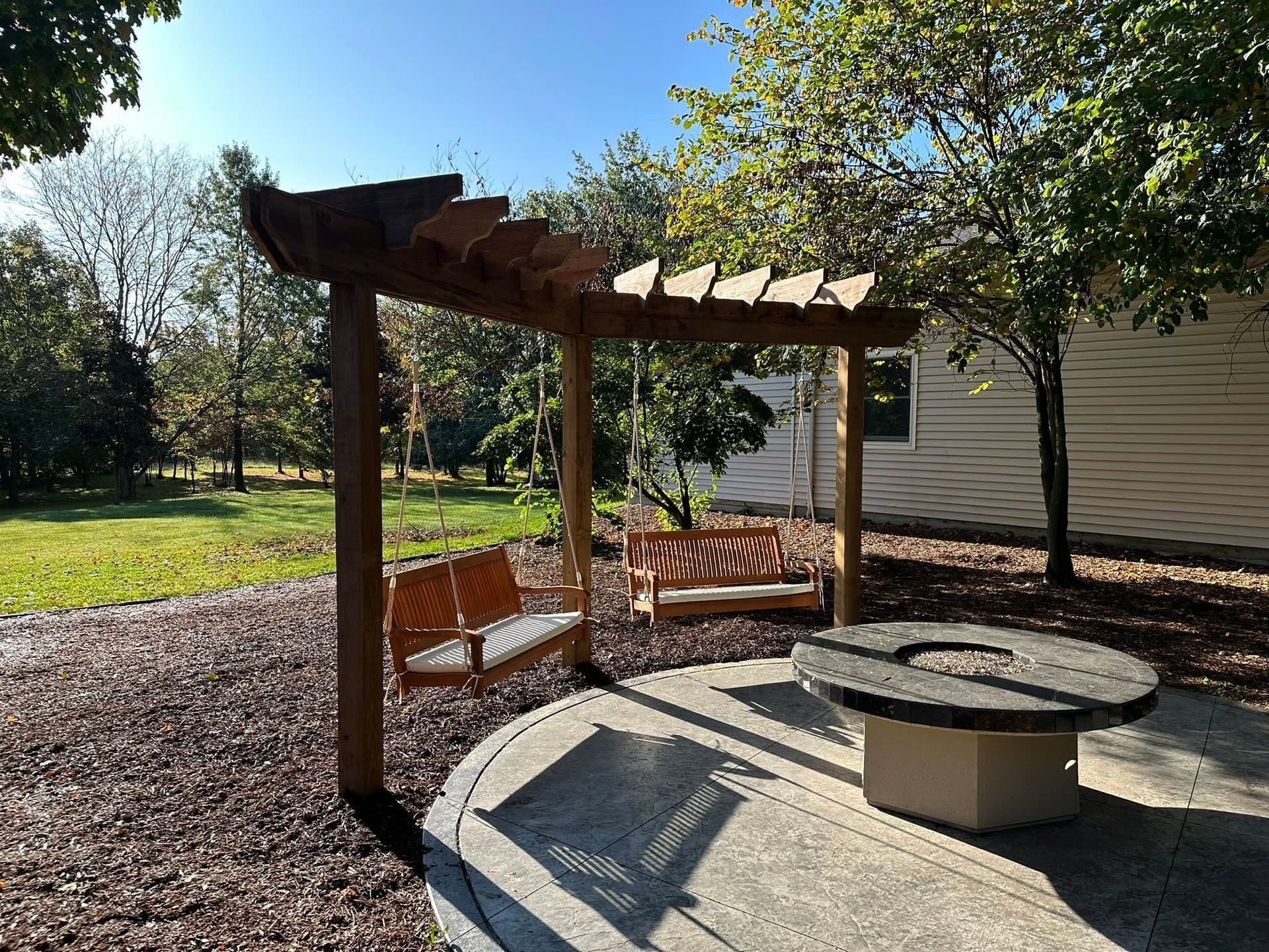 Wooden pergola with bench seating and a round fire pit in a sunny backyard garden