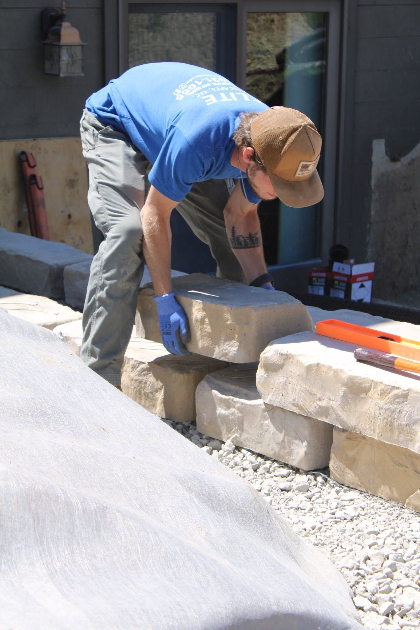 Worker in blue shirt and cap lifting a stone block onto a low wall outdoors.