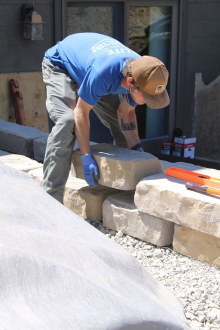 Worker in blue shirt and cap laying a stone block on a low wall outdoors