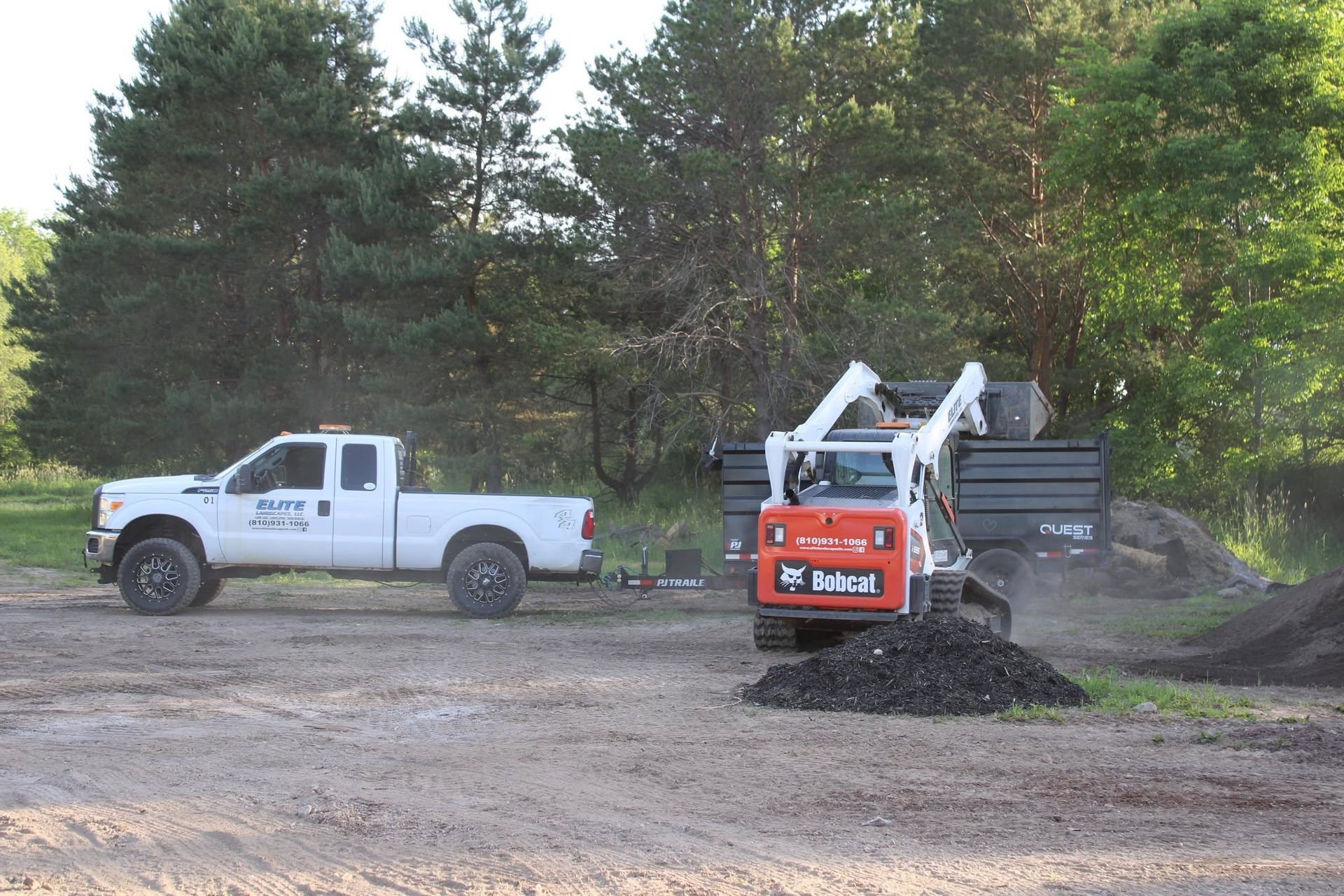 White pickup truck beside a compact orange excavator moving dirt on a wooded worksite