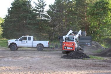 White pickup truck beside a compact orange excavator moving dirt on a wooded worksite