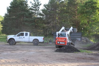 White pickup truck beside a compact orange excavator moving dirt on a wooded worksite