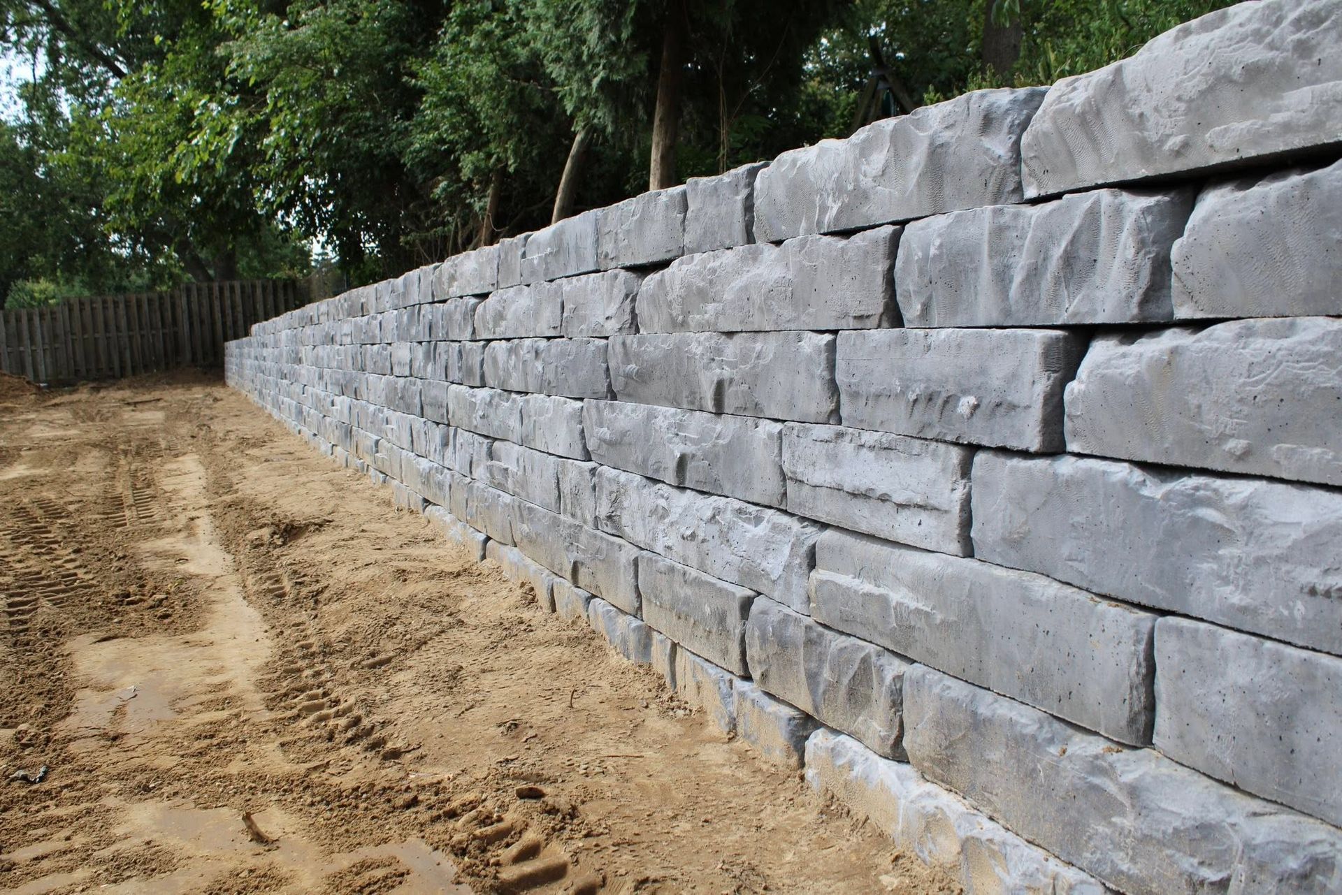 Stone retaining wall beside a sandy path, with trees and a wooden fence in the background.