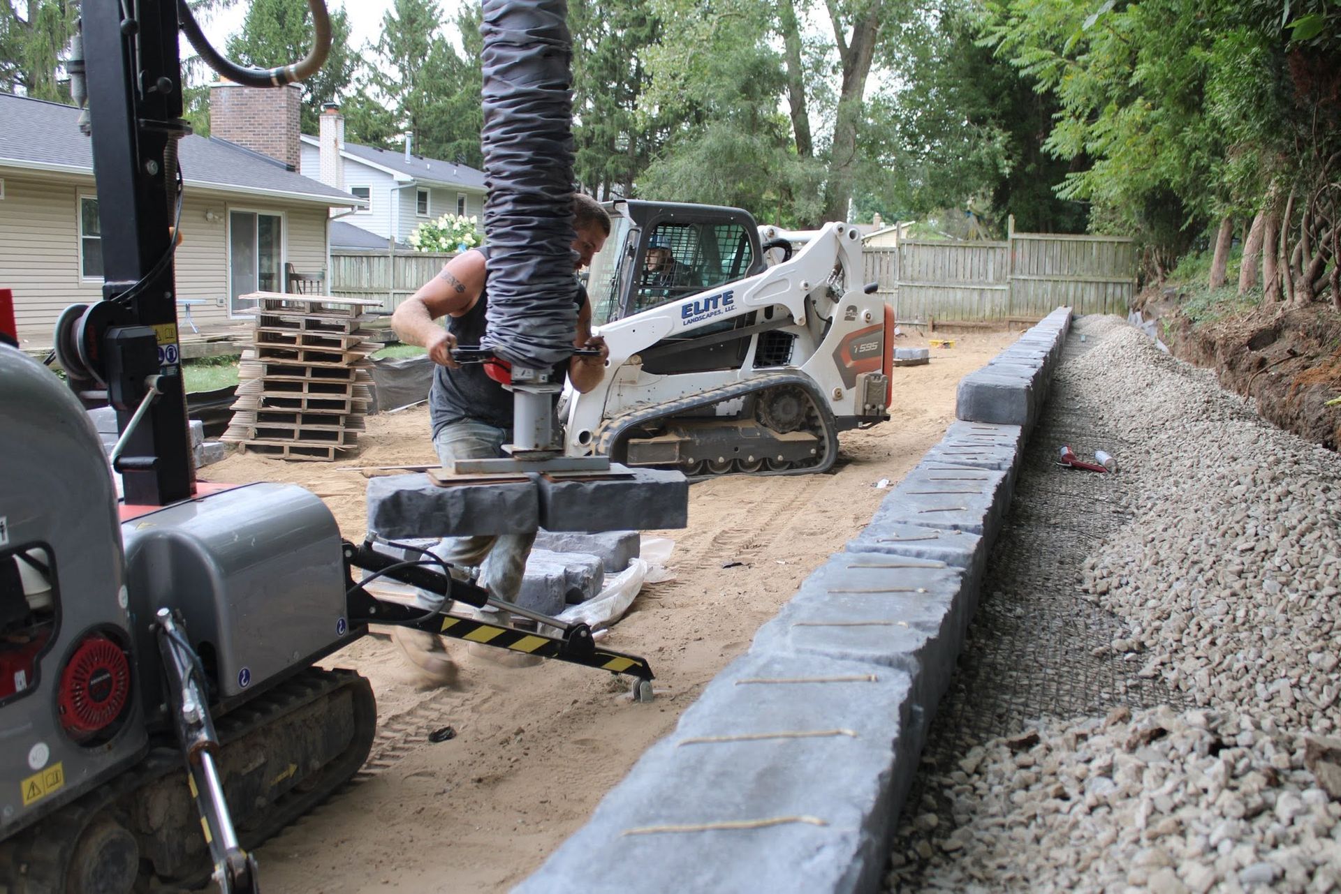Construction equipment at a curbside worksite, with a trench lined by concrete edging and a skid steer nearby.