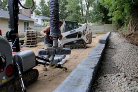 Stone retaining wall beside a sandy path, with trees and a wooden fence in the background.