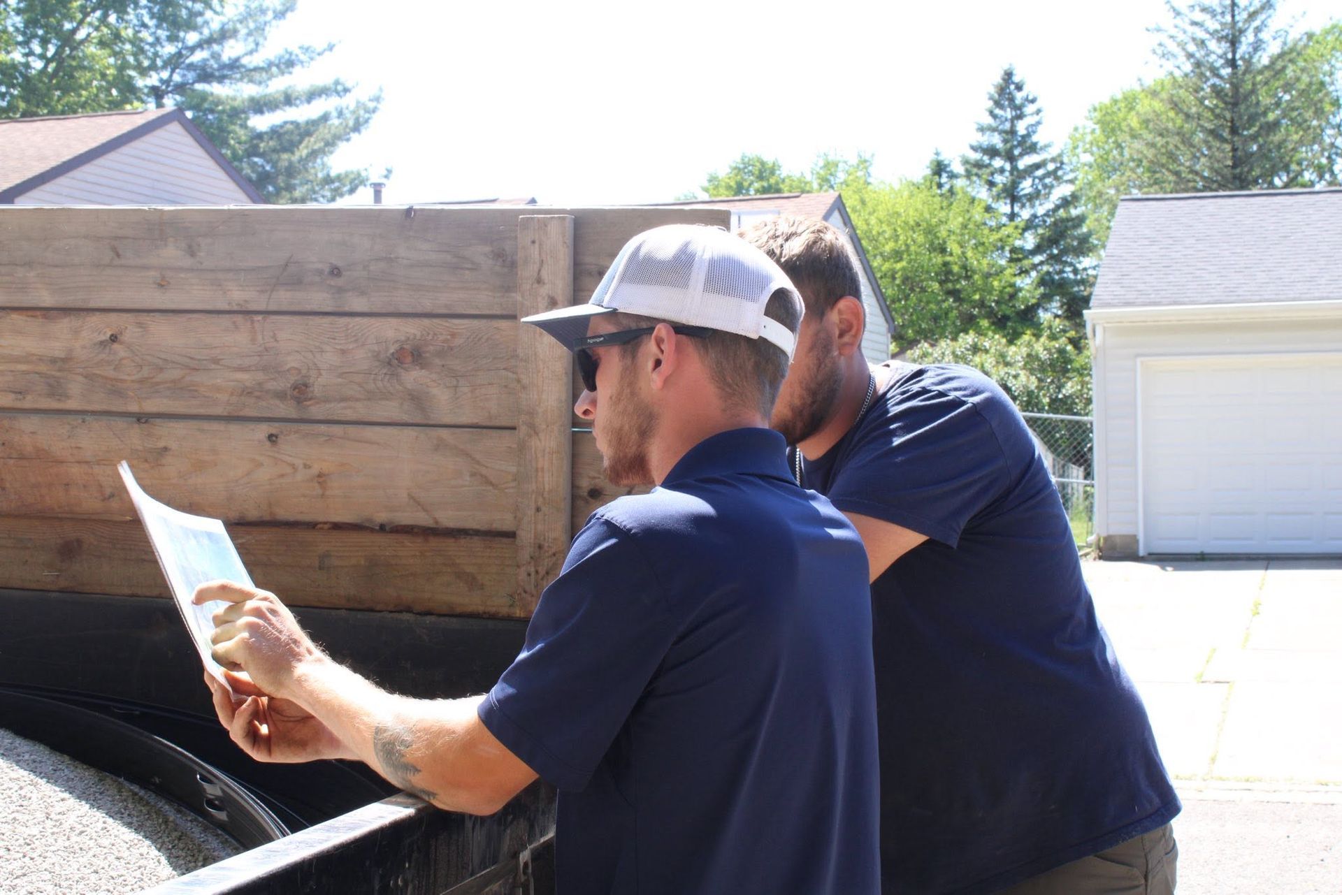 Two workers in navy shirts and caps inspect a wooden dump trailer outdoors.