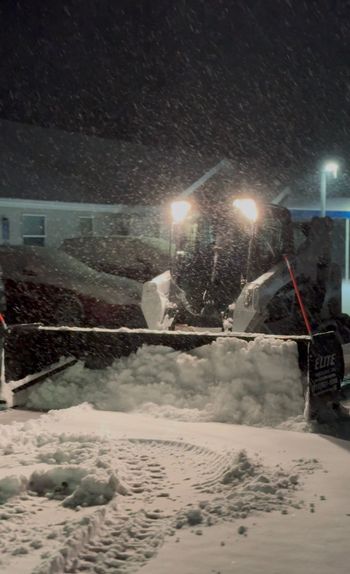 Snowplow clearing a snowy road at night during heavy snowfall, bright lights shining through the snow.
