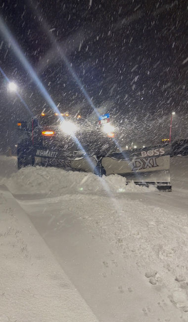 Snowplow clearing a snowy road at night during heavy snowfall, bright lights shining through the snow.