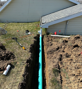 Trenched yard with black drain pipes and white conduit running through grass beside a house.