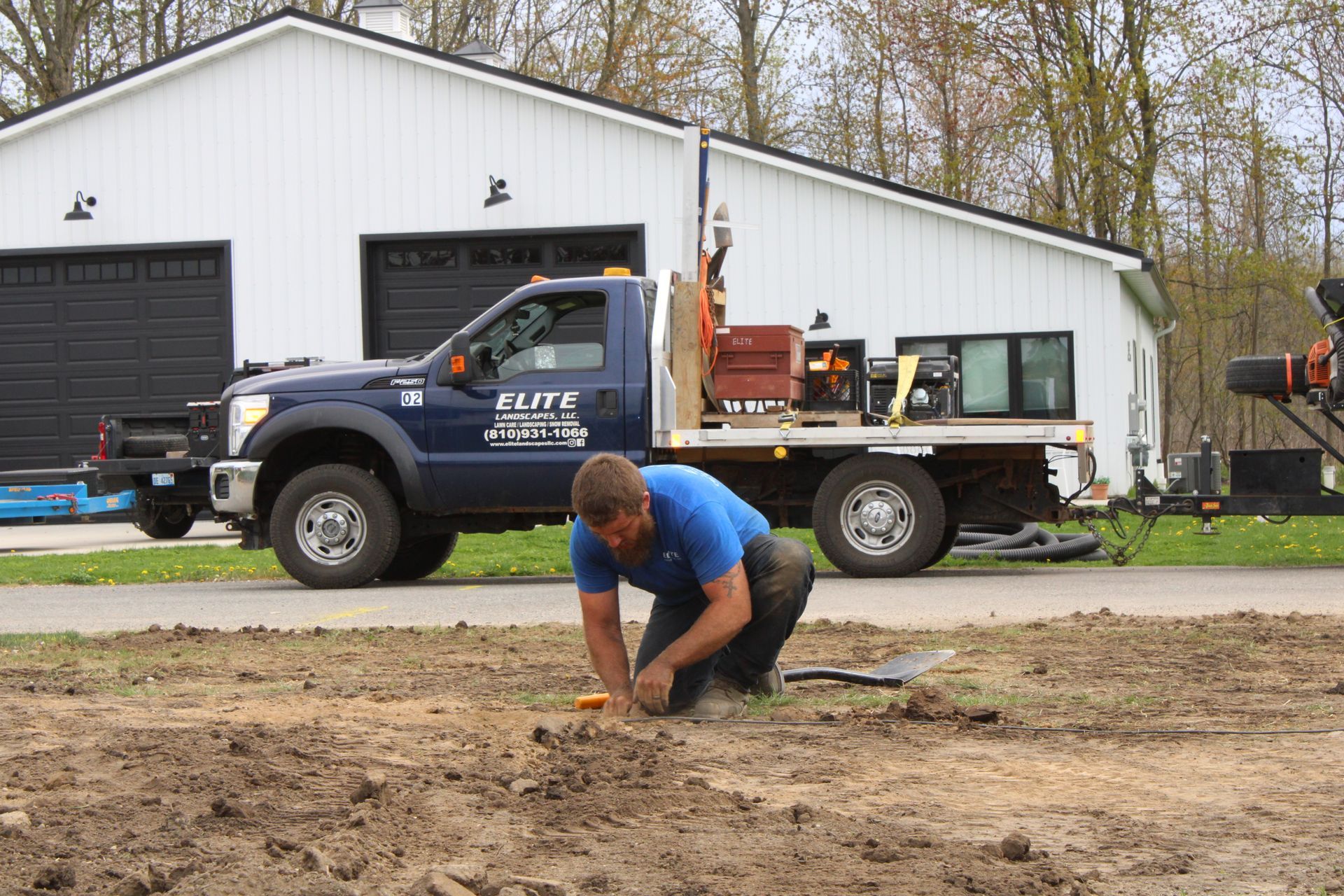 Worker kneeling in dirt beside a flatbed truck parked in front of a white garage