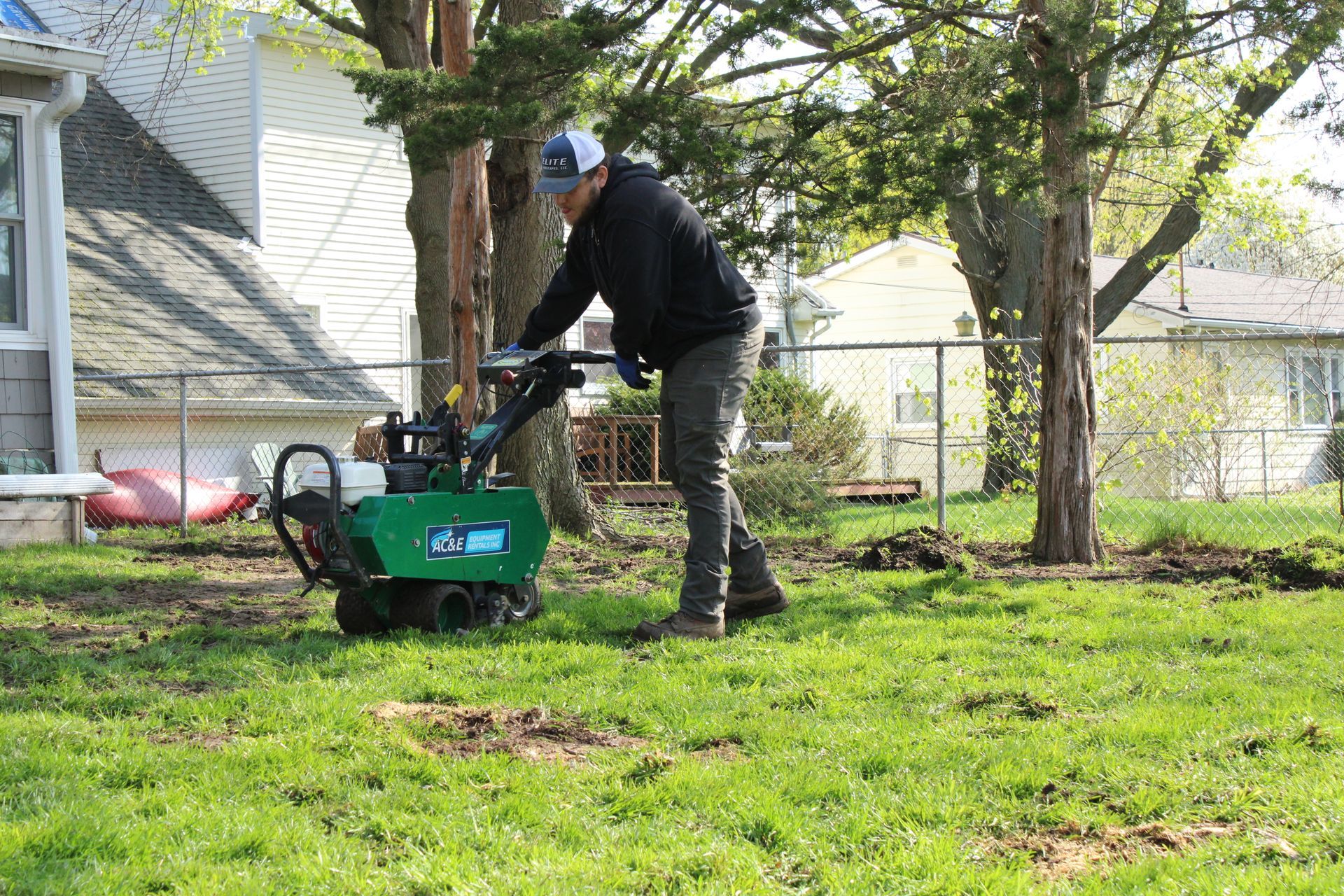 Person using a green tiller in a grassy backyard near trees and a house