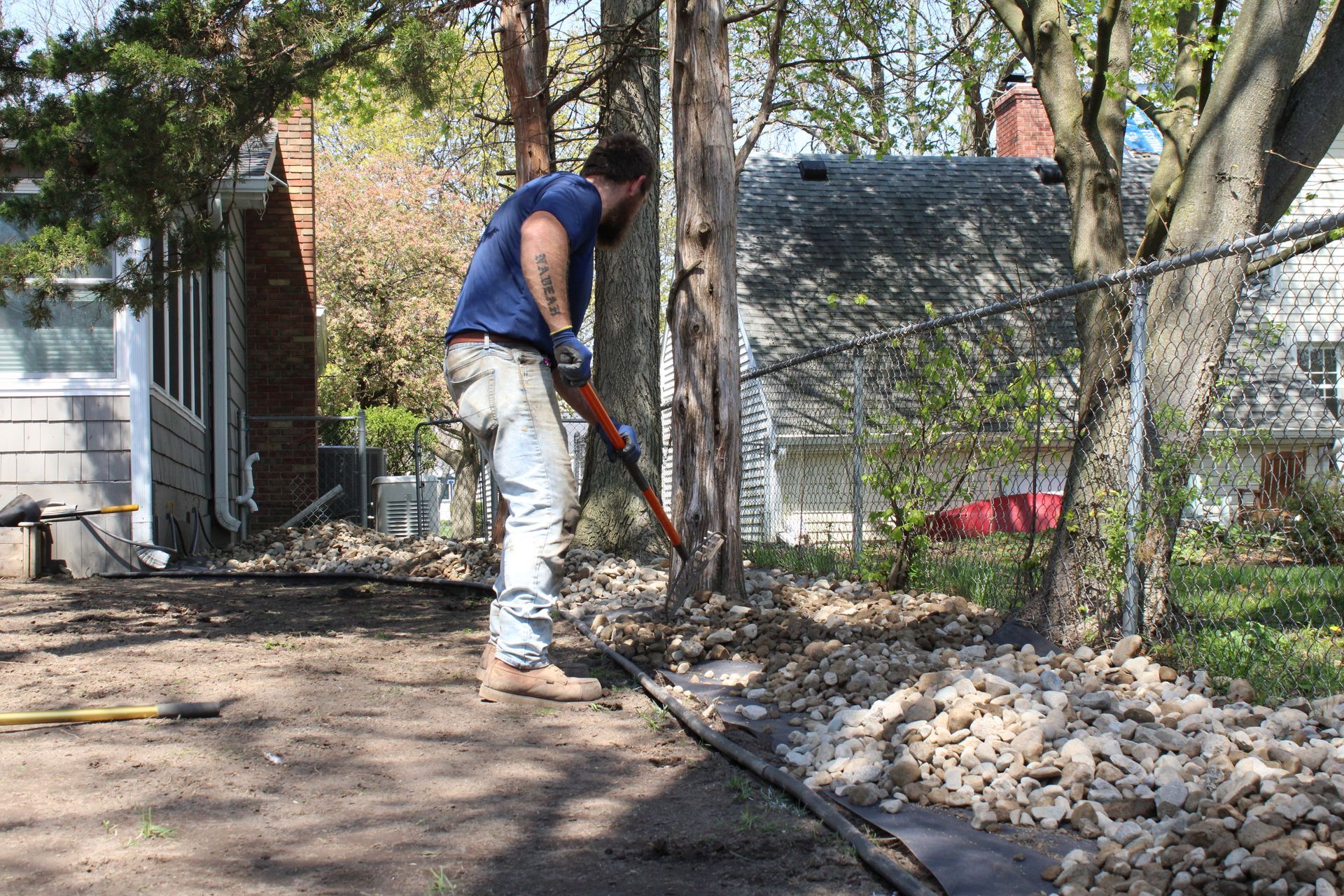 Person using a shovel to clear debris in a backyard beside a chain-link fence and trees