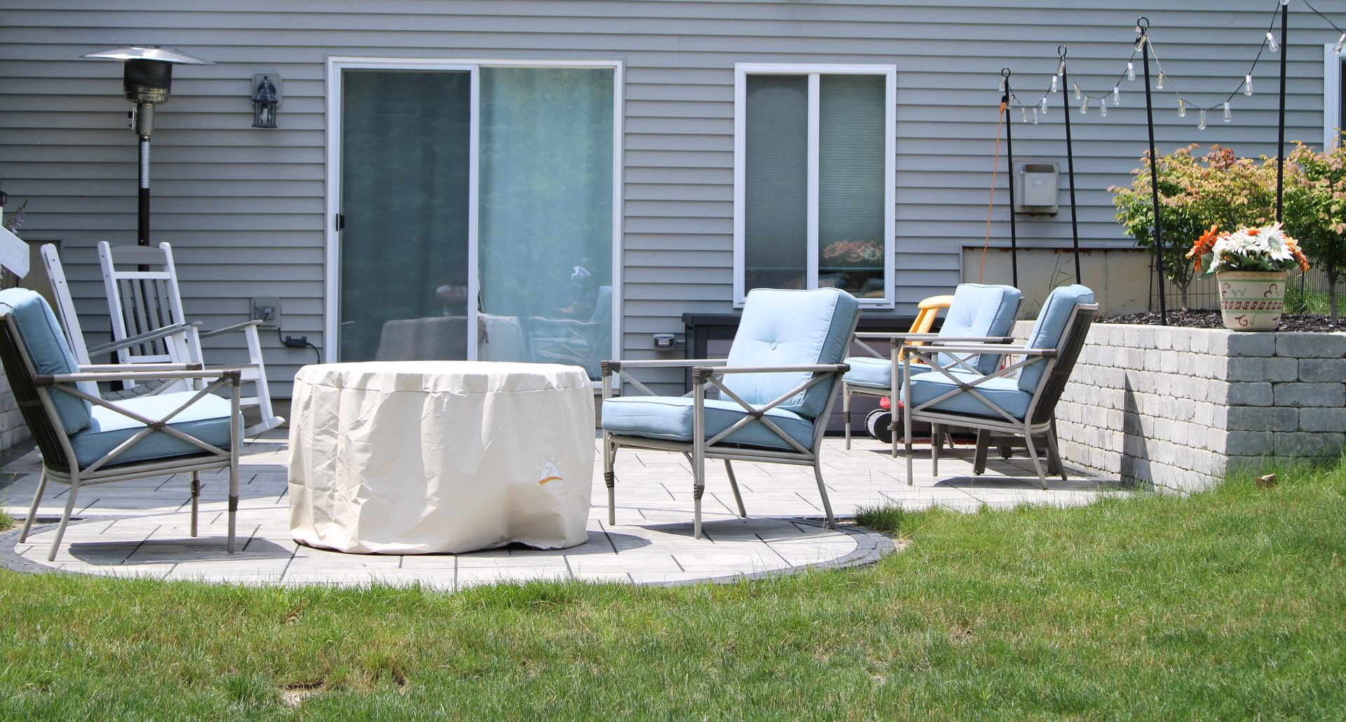 Patio with chairs around a round table on a small lawn beside a house, with potted flowers and sliding doors.
