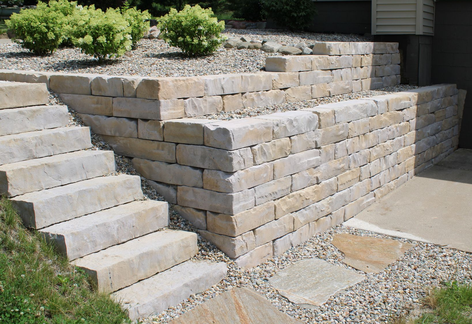Stone retaining wall and concrete steps beside a gravel yard and shrubs in a residential landscape