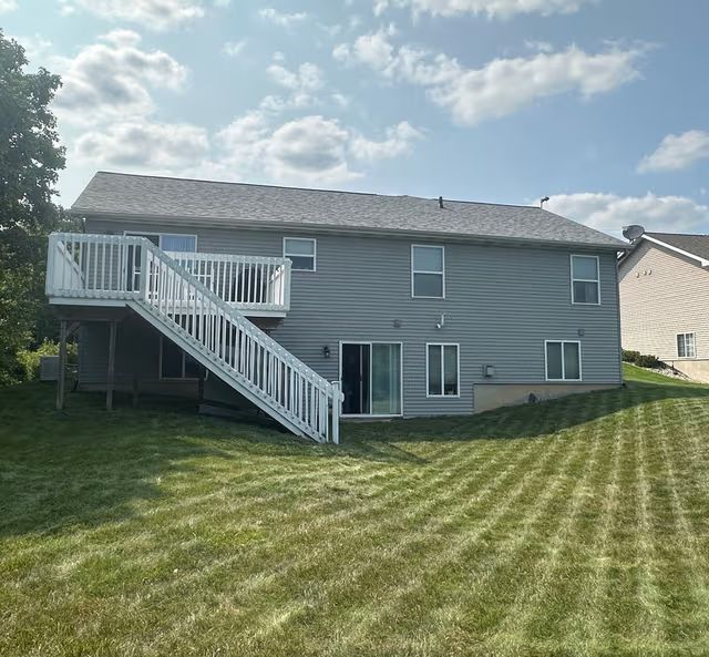 Gray two-story house with white deck stairs, sliding door, and a striped green lawn under a cloudy sky