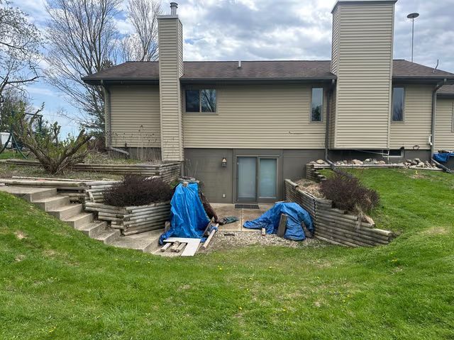 Backyard of a beige house with retaining walls, steps, blue tarps, and a grassy lawn