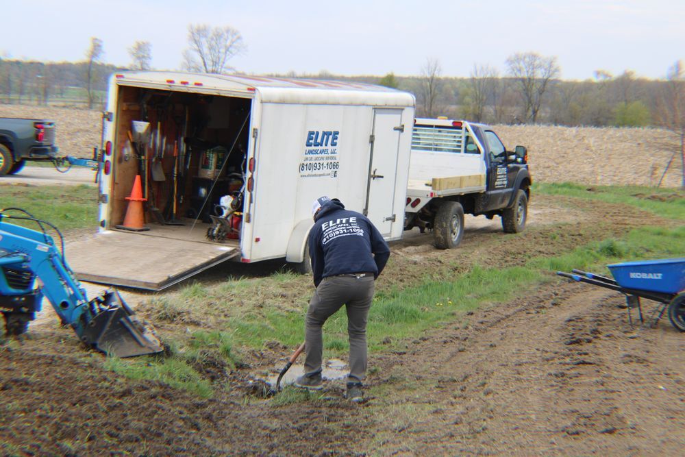Worker in muddy field beside utility truck and equipment trailer, loading gear near a blue excavator