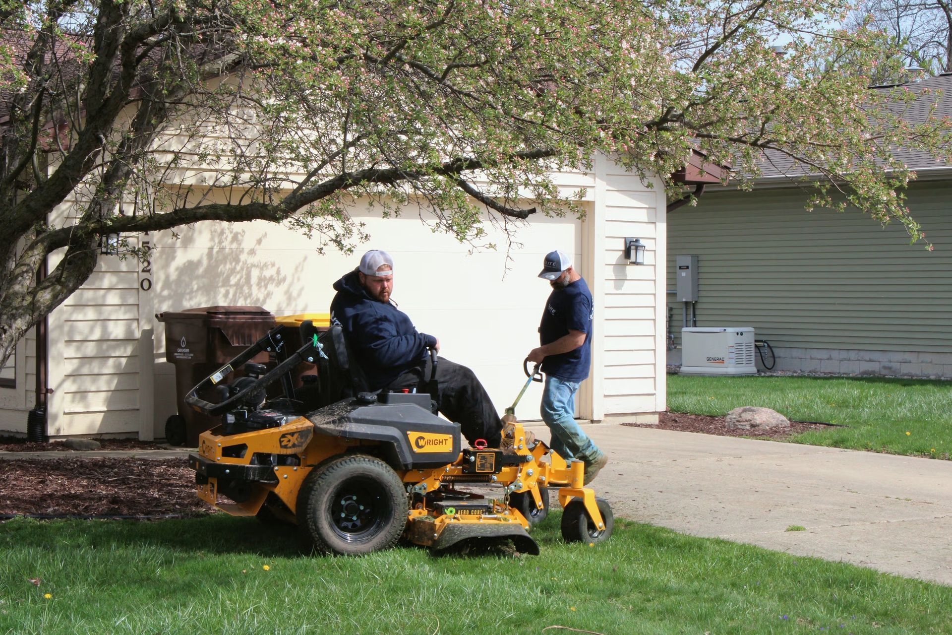 Two people stand by a yellow lawn mower near a driveway in front of a white house.