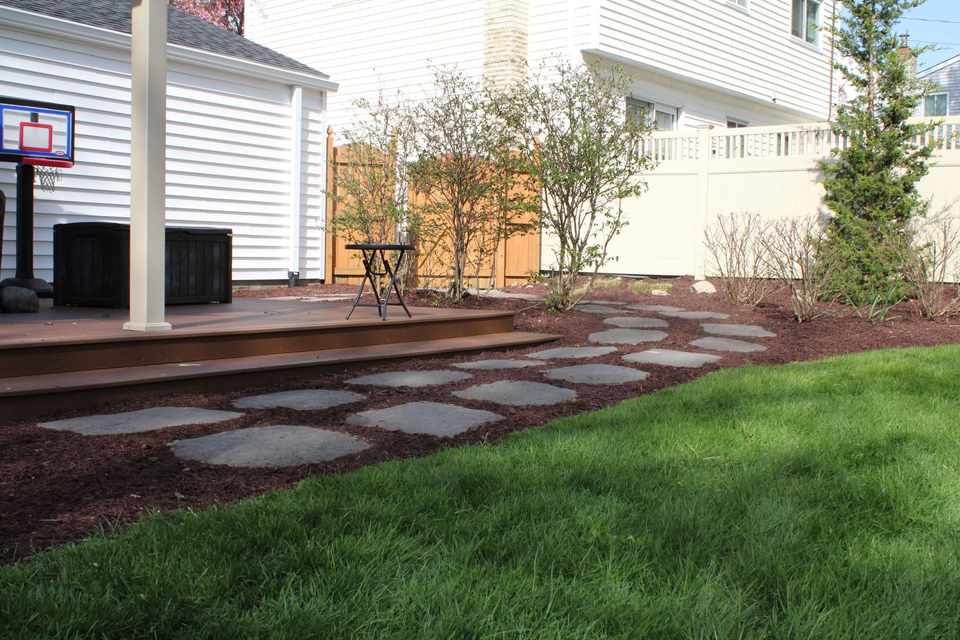 Backyard patio with stone stepping path, mulch beds, lawn, and a small basketball hoop near a house