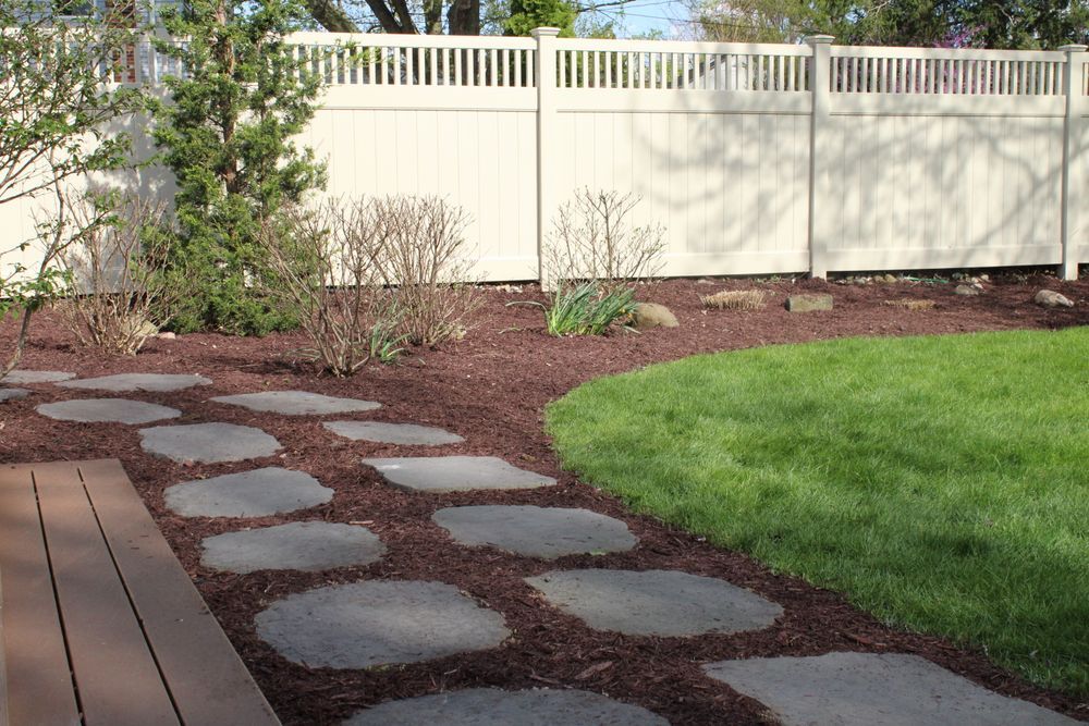 Backyard garden path with stepping stones, wooden deck, green lawn, and white fence