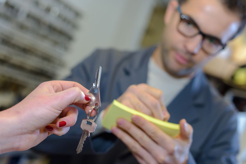 Hand Holding House Keys Toward a Blurred Person Writing on A Yellow Notepad — Terrace Locksmiths in Newcastle, NSW
