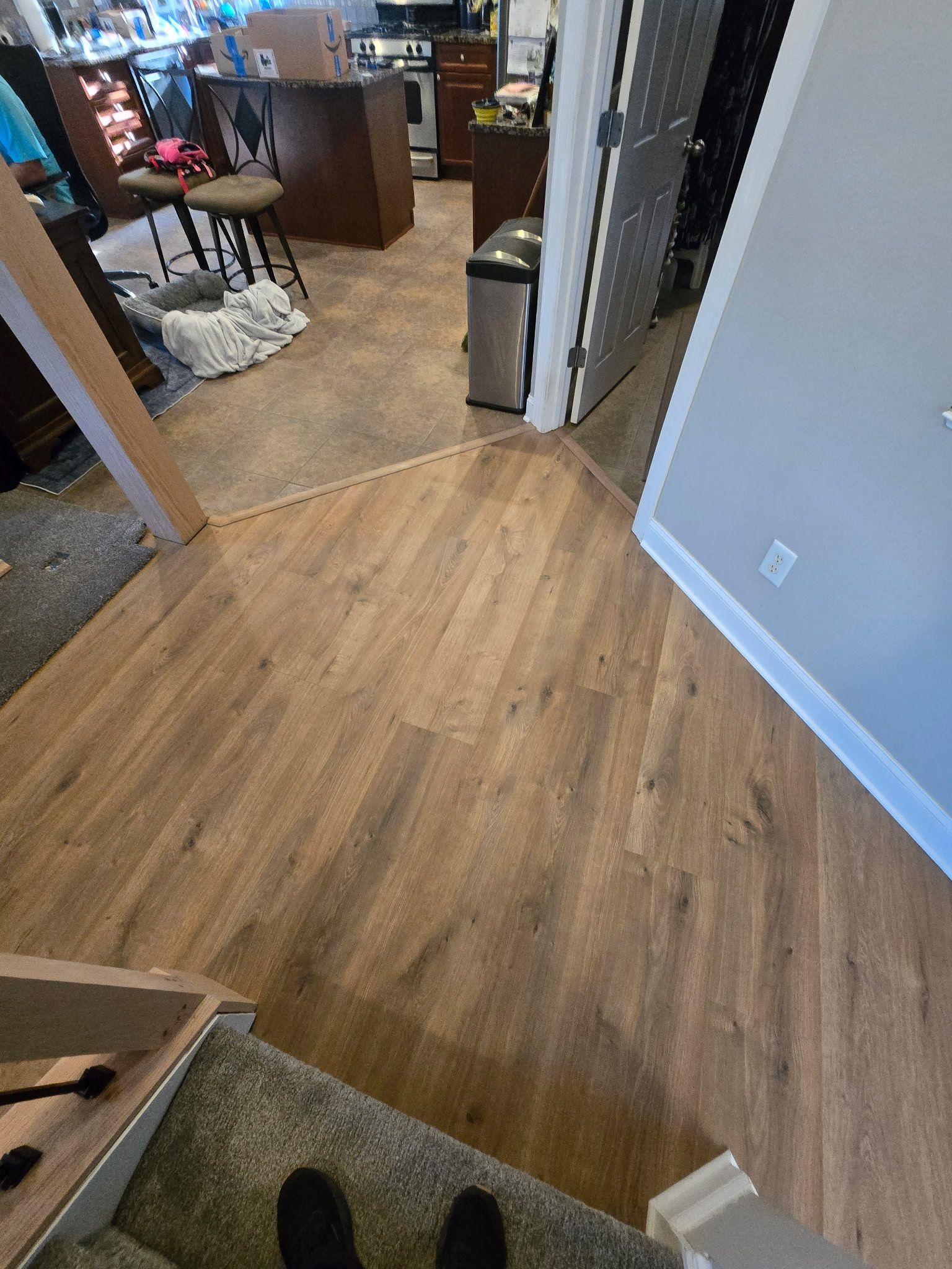 Looking down at brown wood flooring transitioning to a darker area with a staircase and dark shoes.