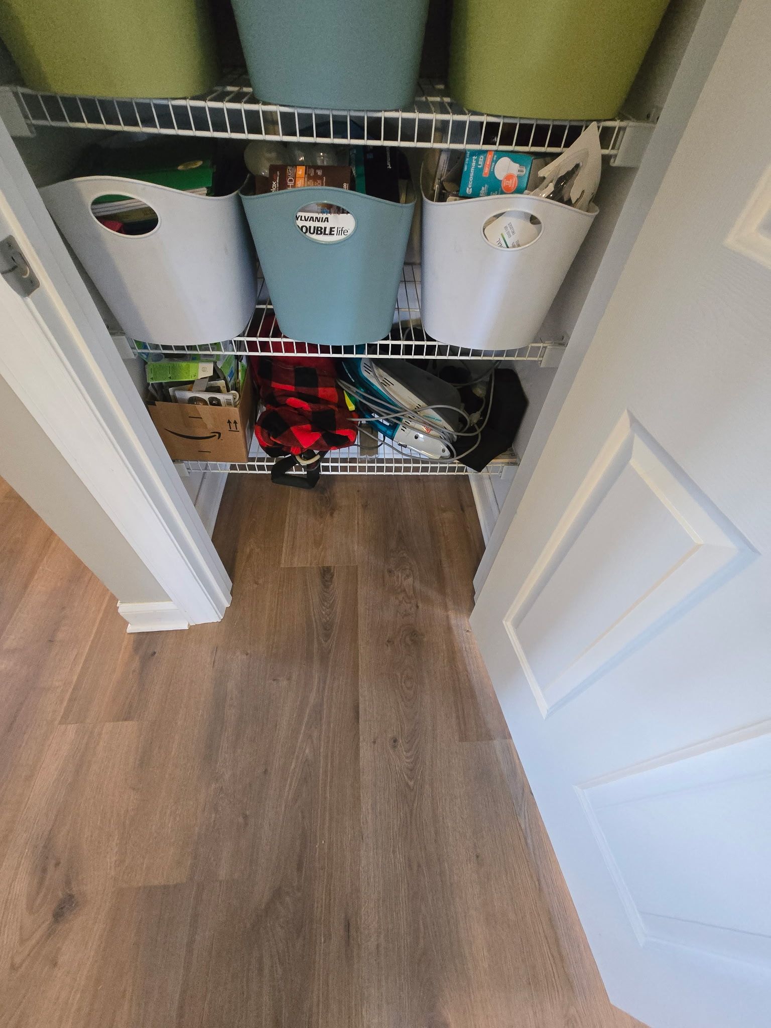 A closet with wire shelves holding various containers in white, blue, and green. The floor is wood-toned.