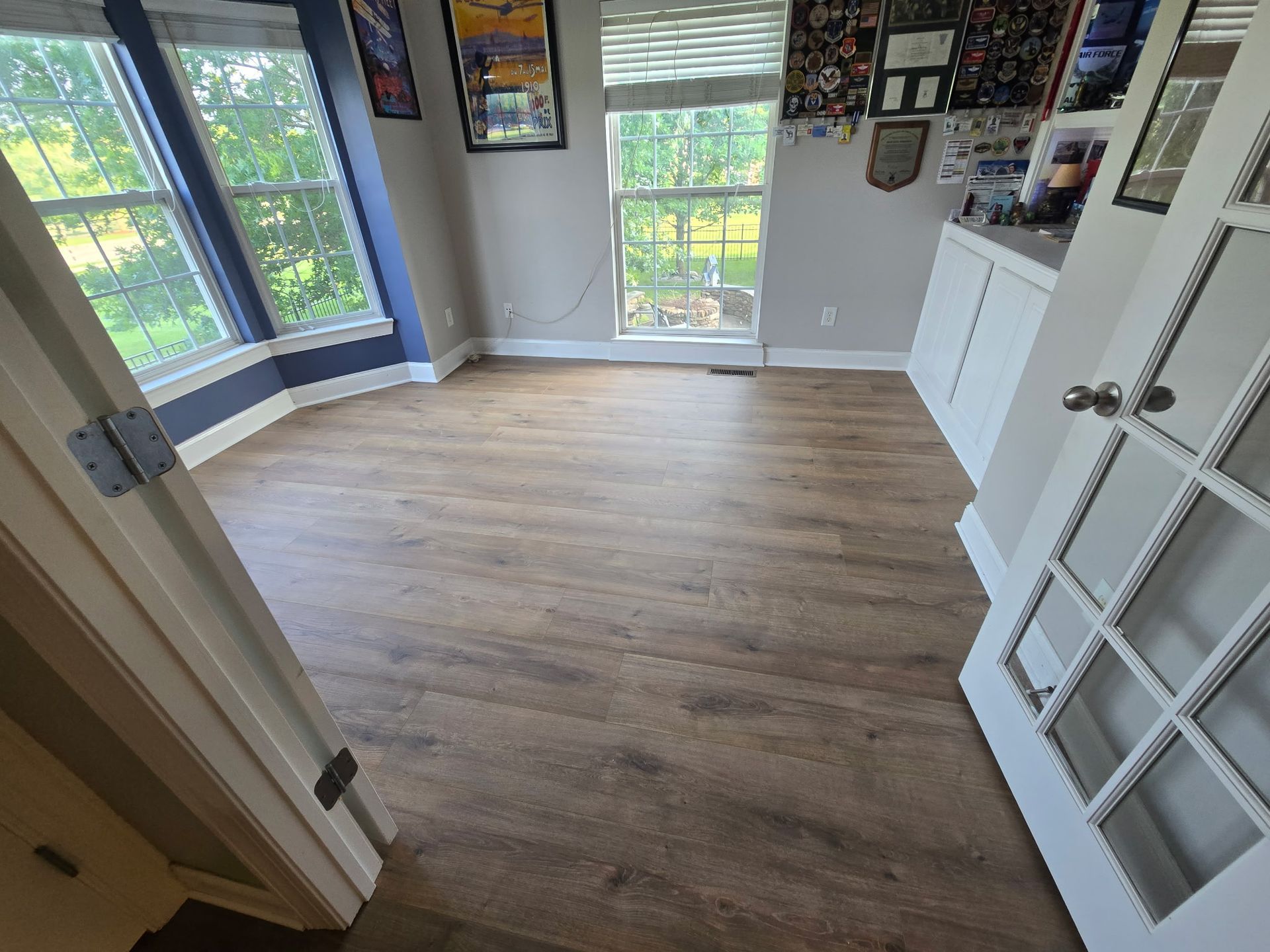 Empty room with wood-look flooring, windows, and a white door; gray walls, some pictures displayed.
