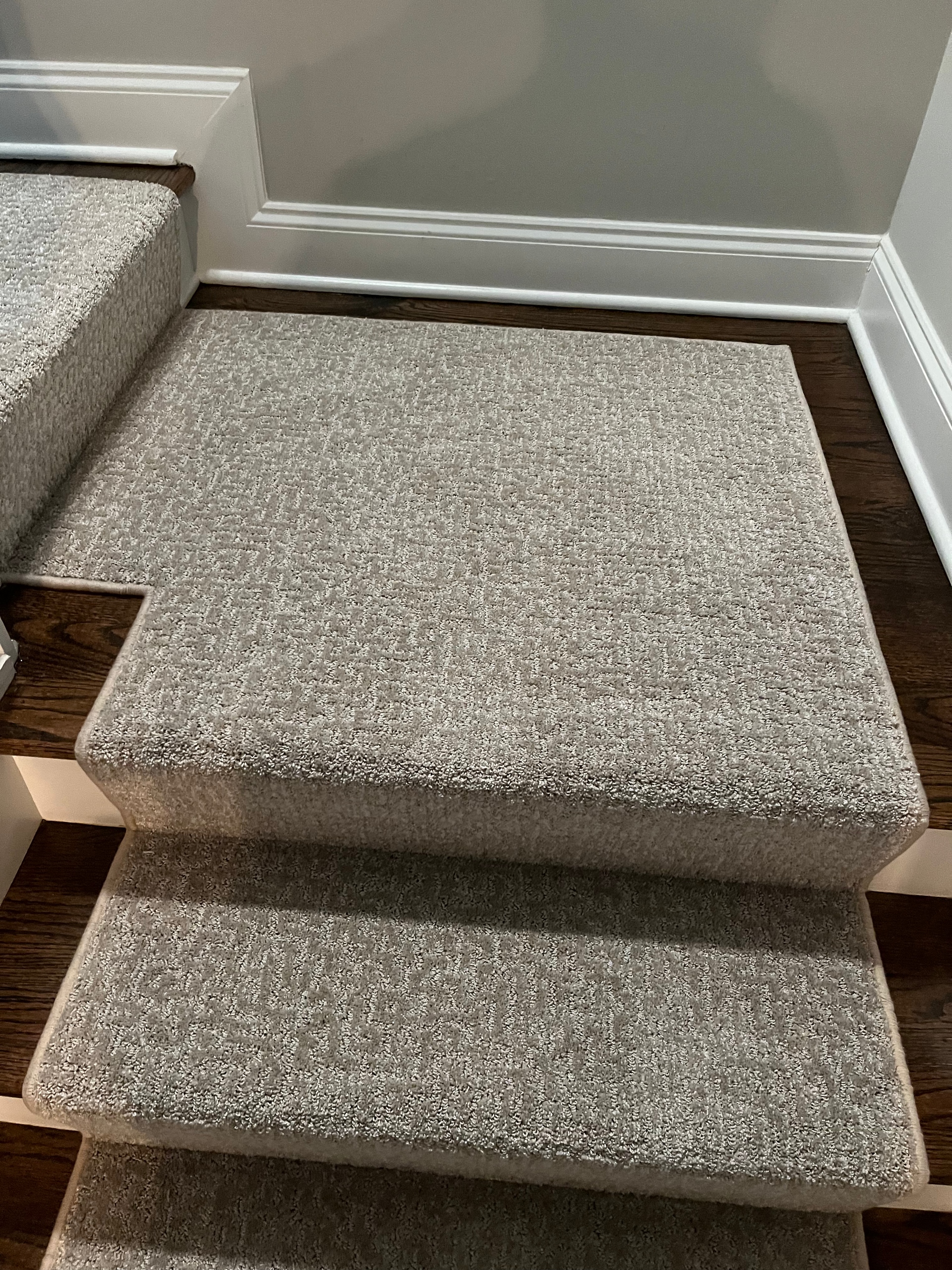 Carpeted staircase with gray and brown tones. The stairs lead upward, against a white-trimmed wall.