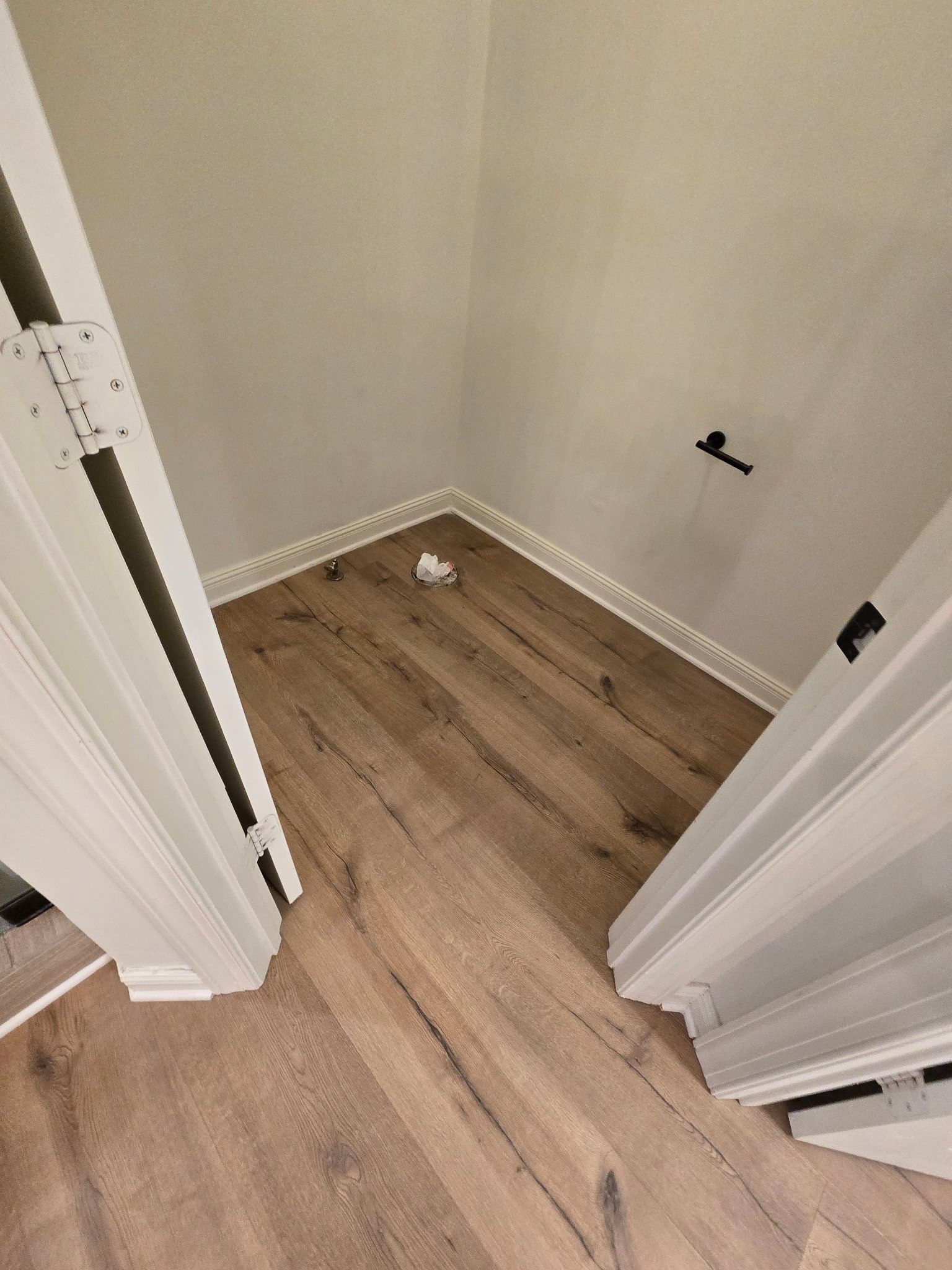 Interior view of a closet corner with wood flooring, white trim, and off-white walls. Two white door frames.